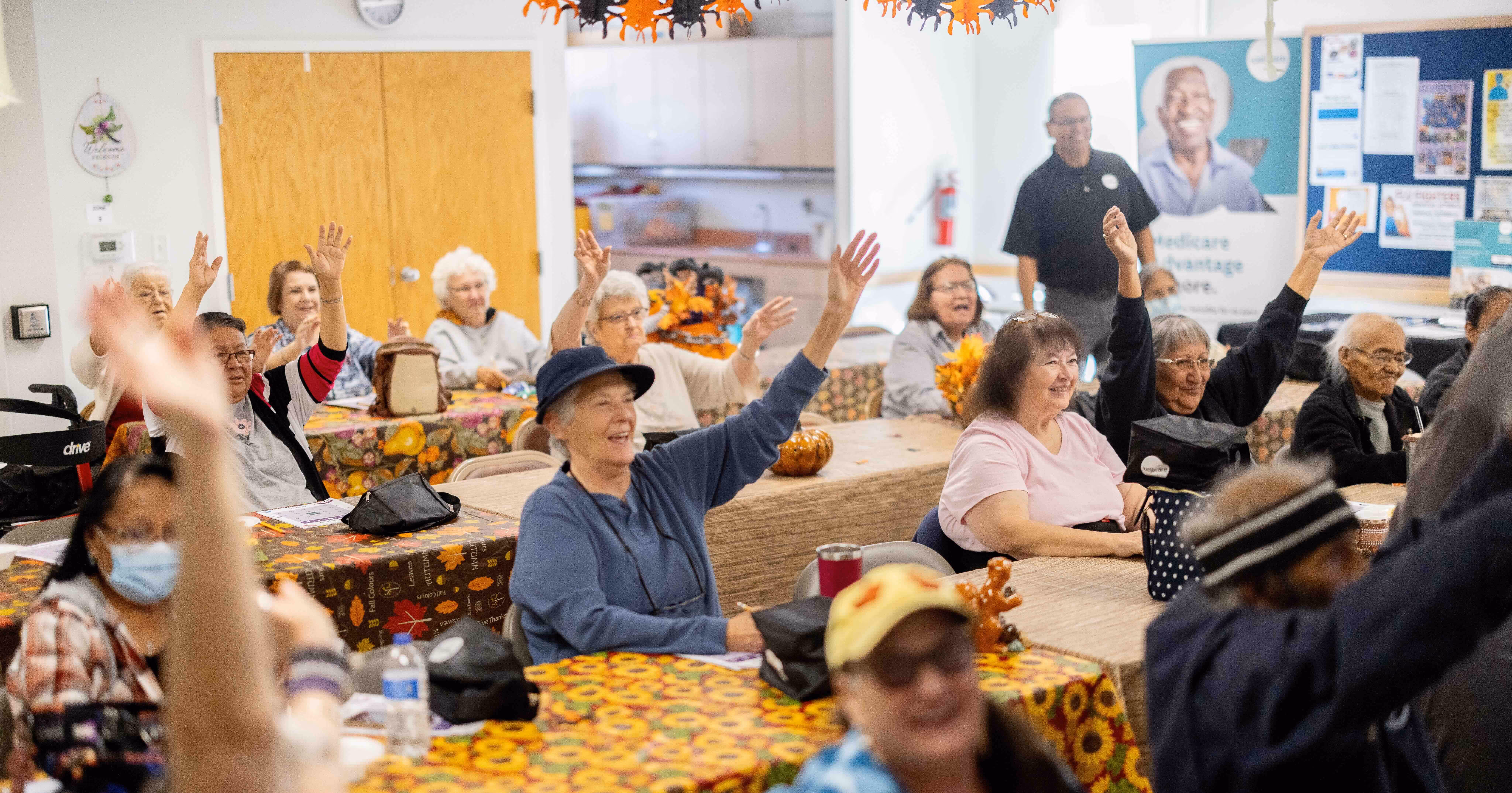 A group of elderly people seated at decorated tables in a community room, many raising their hands and smiling.