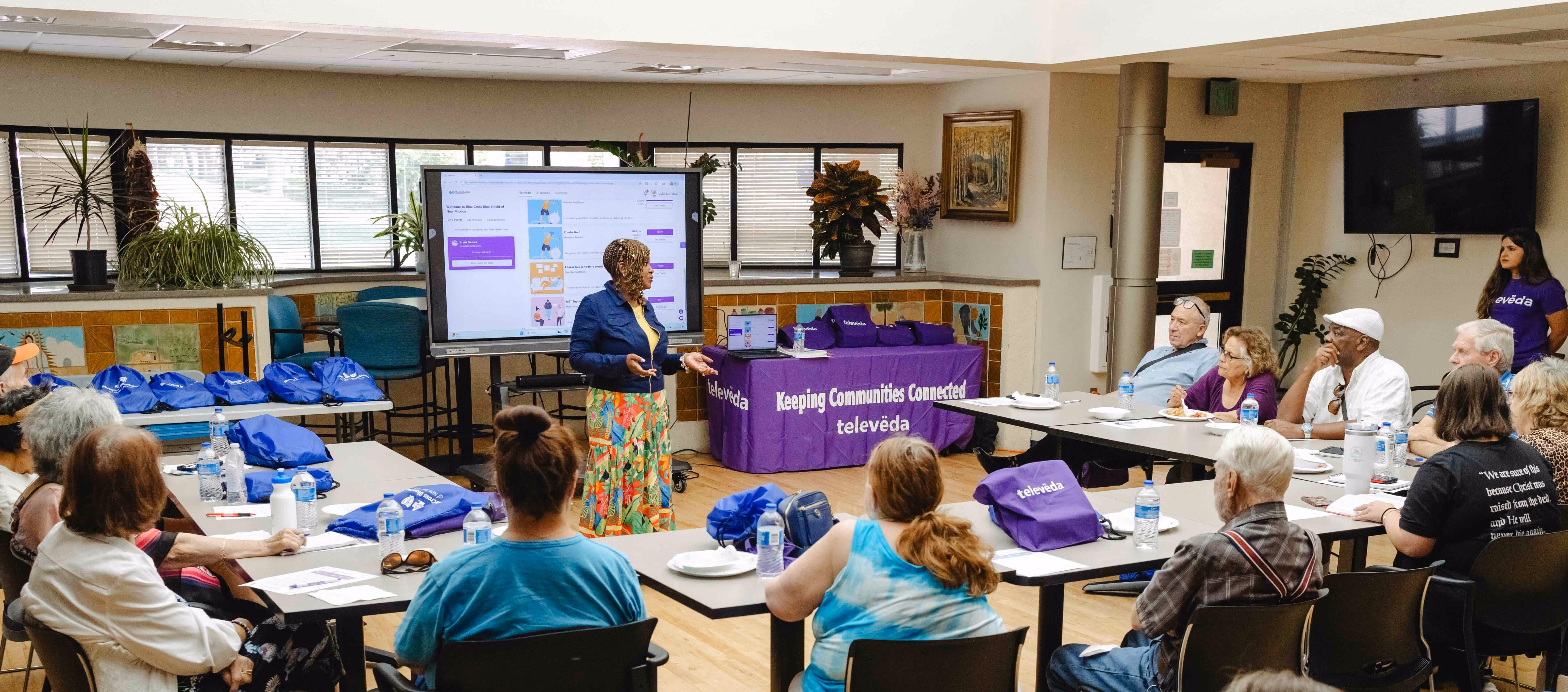 Woman presenting a community workshop to a diverse group of seated adults in a meeting room with 'televeda' branded materials.
