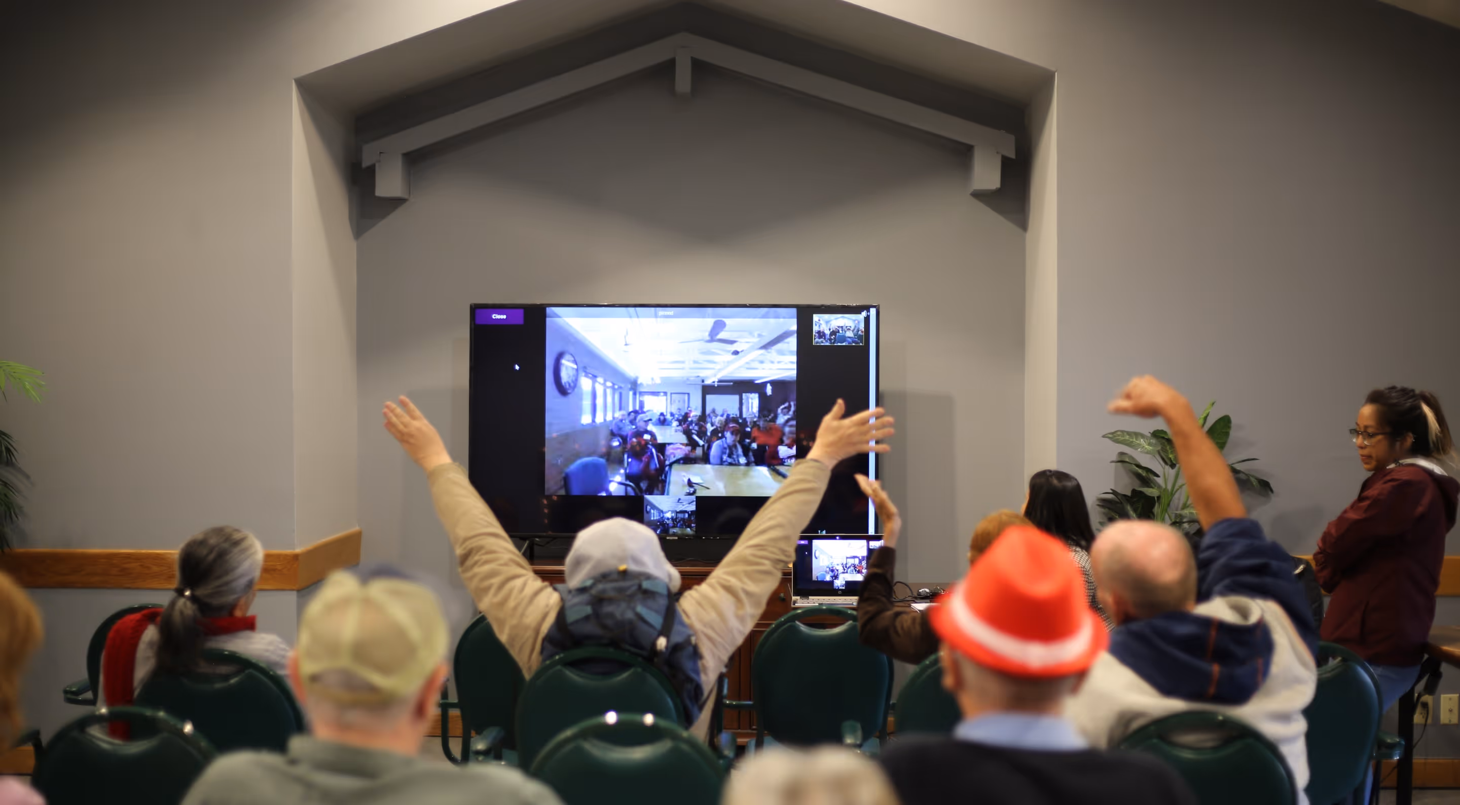 Group of people seated in a room watching a large screen showing a video call with another group, some raising their arms in greeting.