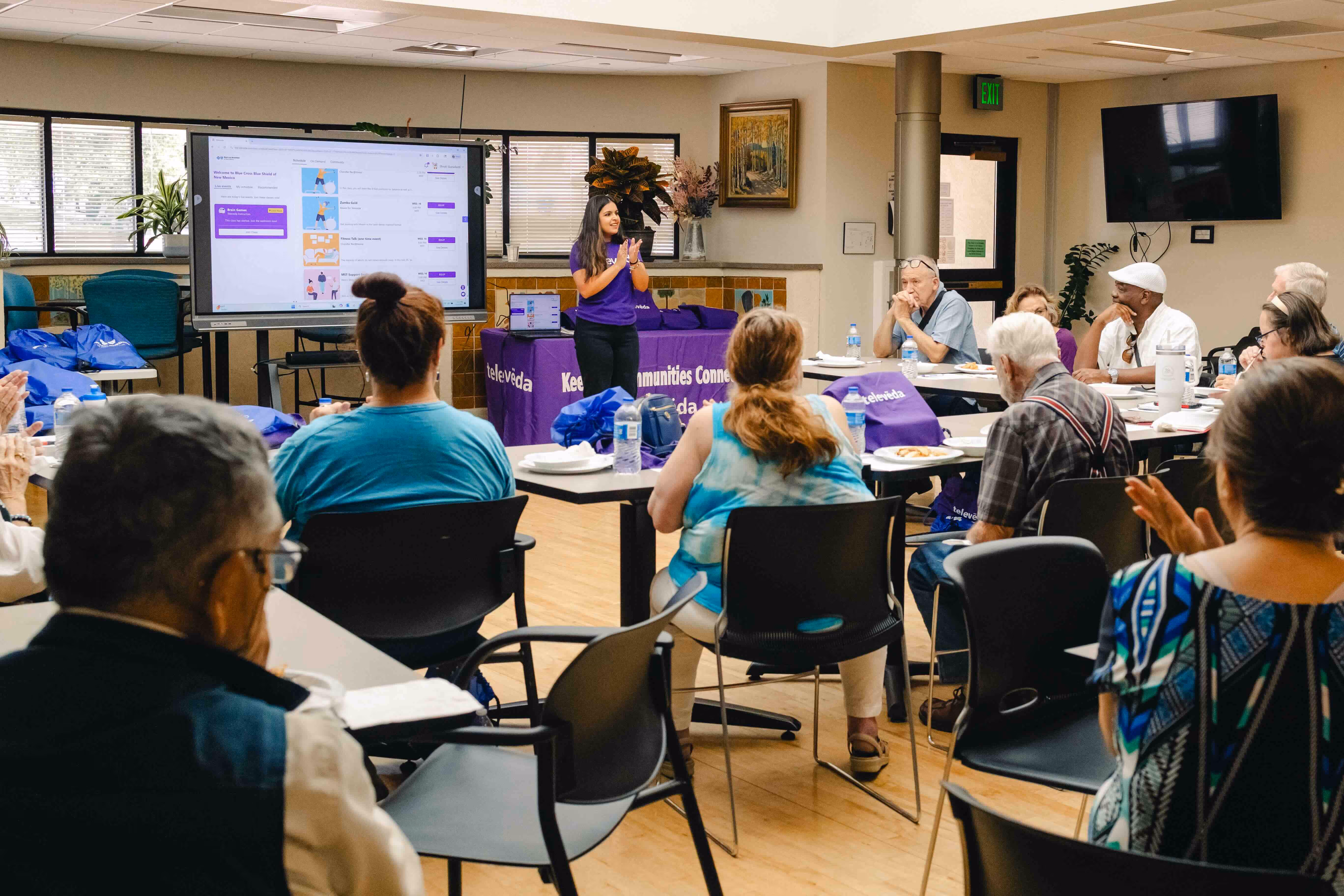 A woman in a purple shirt stands and claps while leading a workshop with seated attendees watching a presentation on a large screen in a community room.