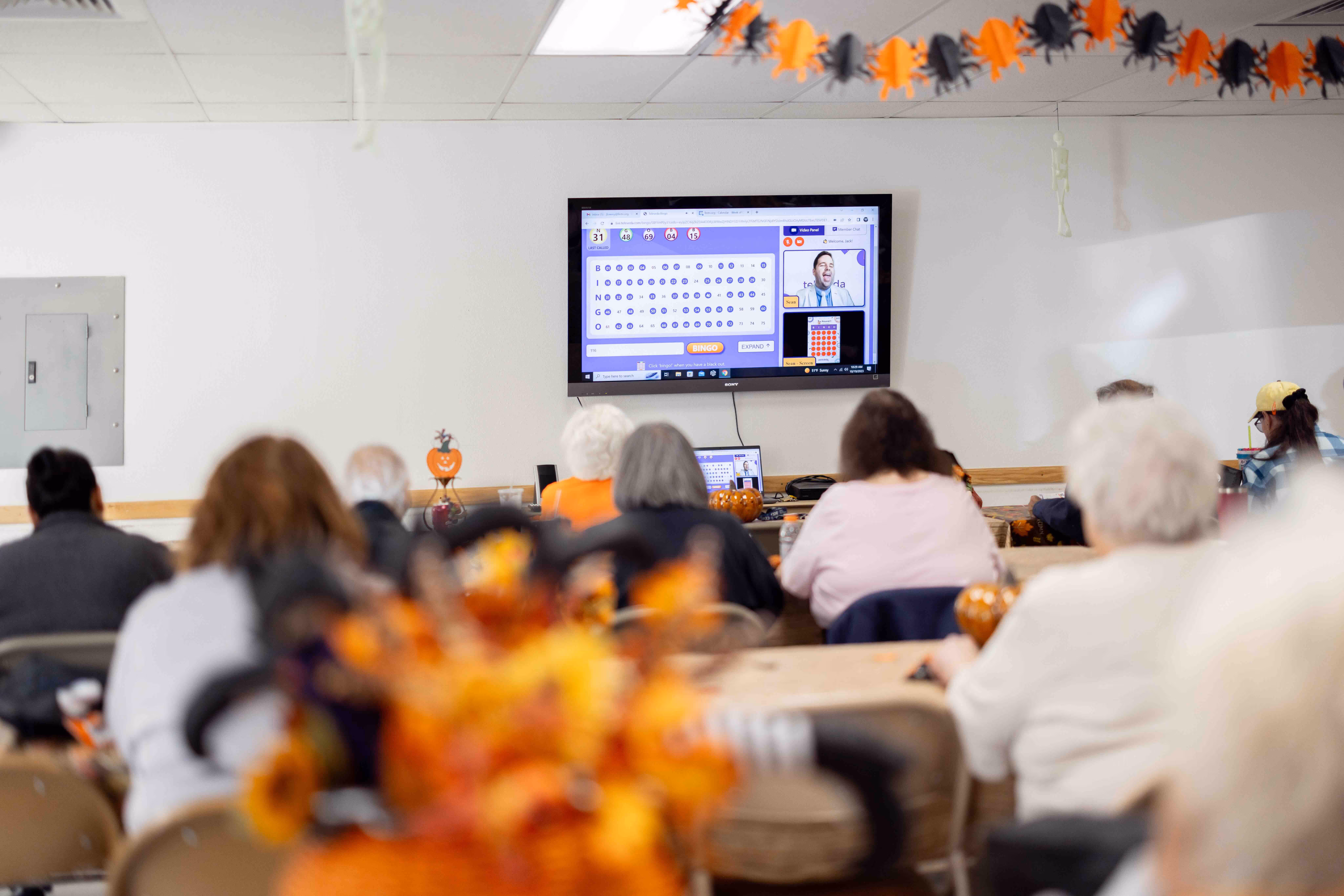 A group of people sitting in a room watching a bingo game on a wall-mounted TV screen, with Halloween decorations visible.