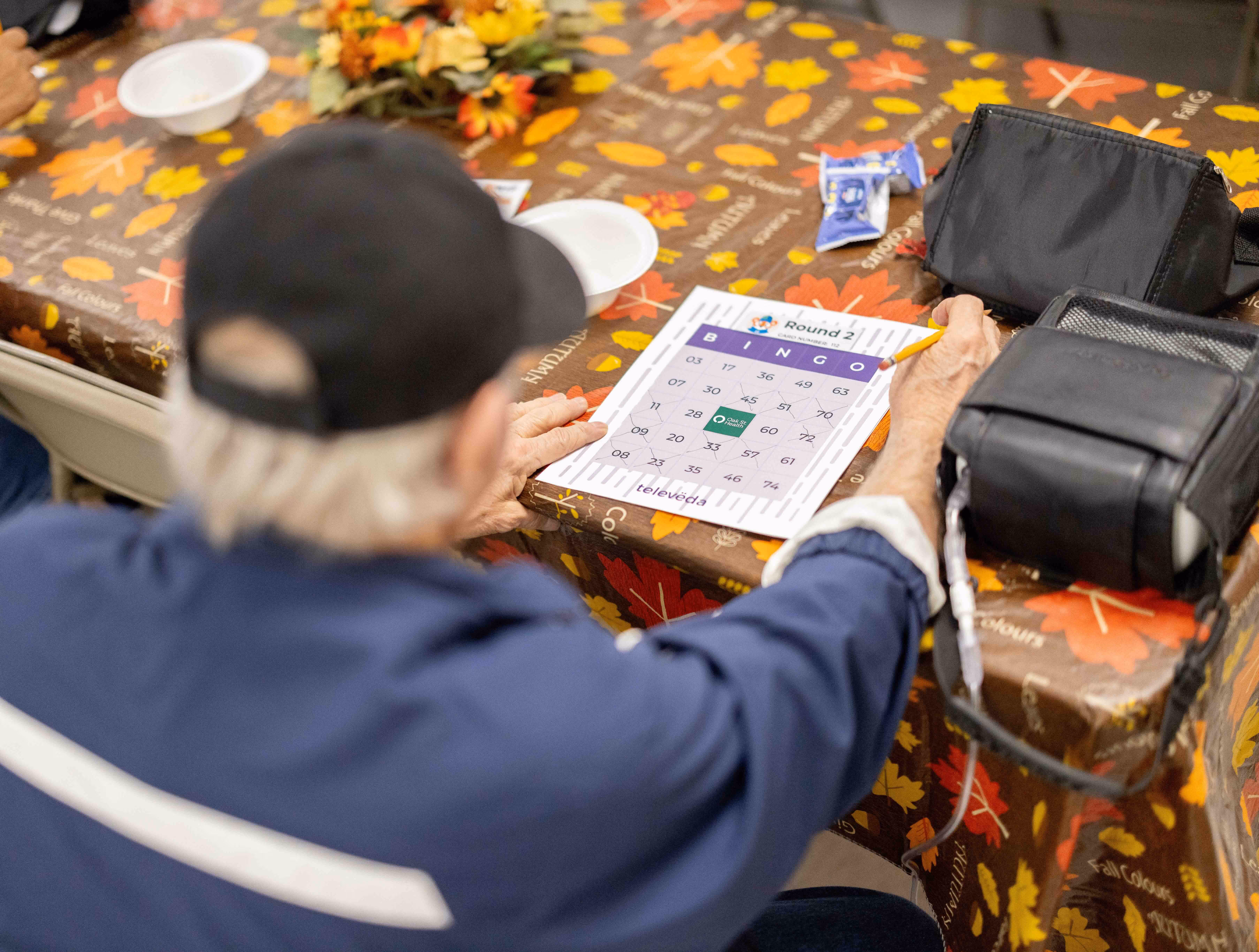 An elderly person wearing a black cap and blue jacket is holding a pencil and playing Bingo at a fall-themed table.