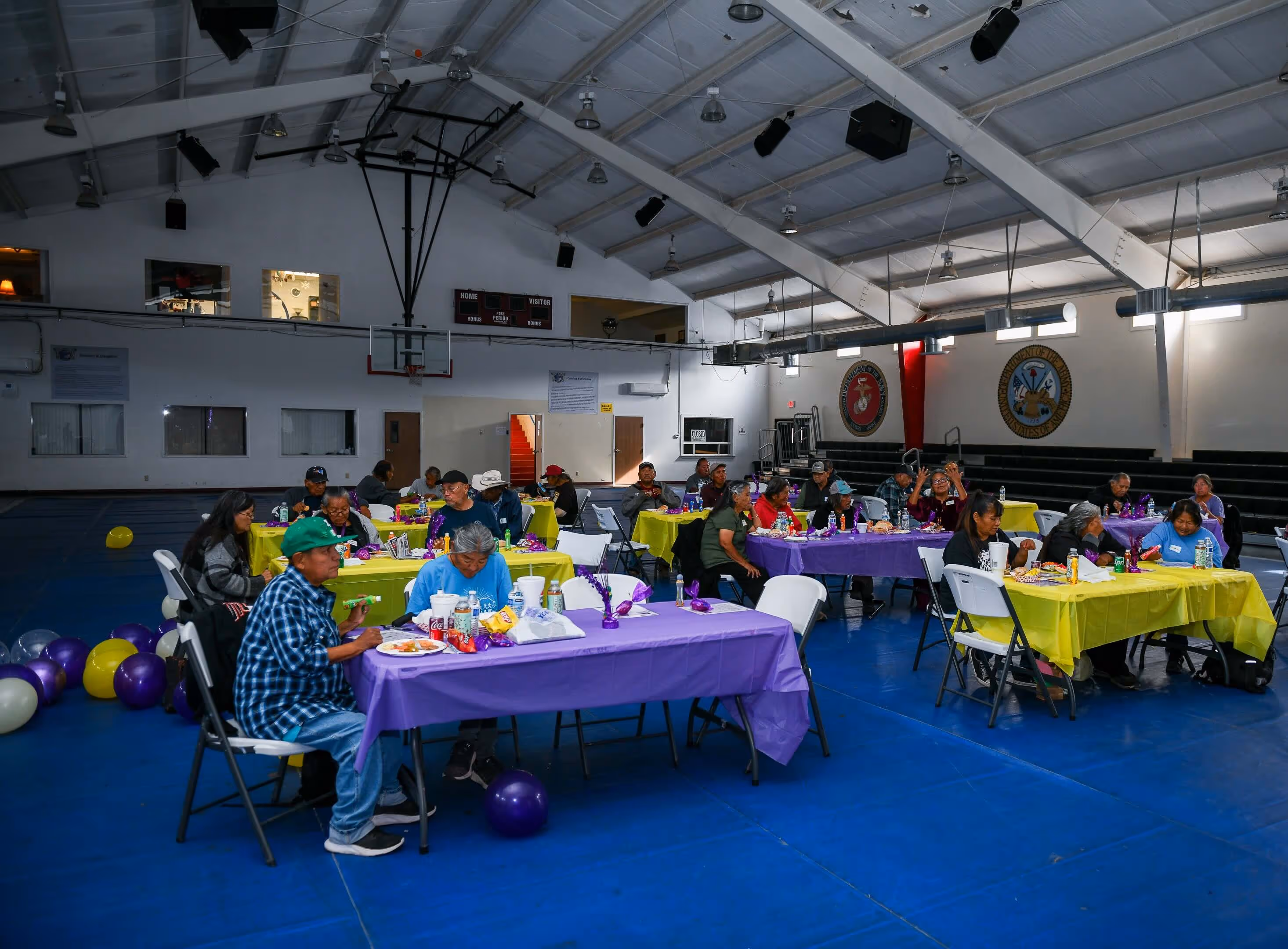 Group of people seated at tables with purple and yellow tablecloths eating and socializing inside a gymnasium decorated with balloons.