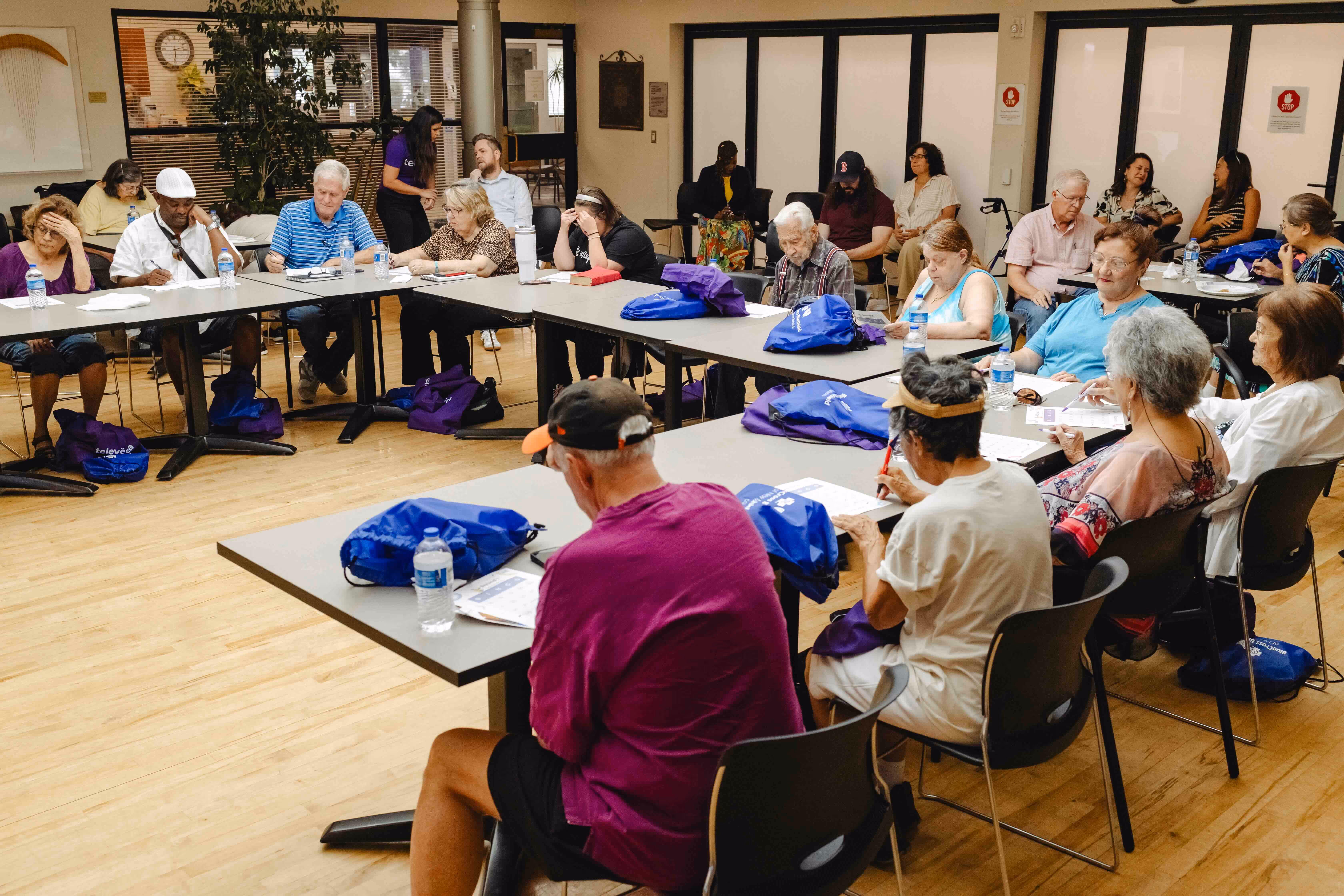 A diverse group of adults seated around tables in a classroom setting, engaged in writing or reading activities with water bottles and blue and purple bags on the tables.