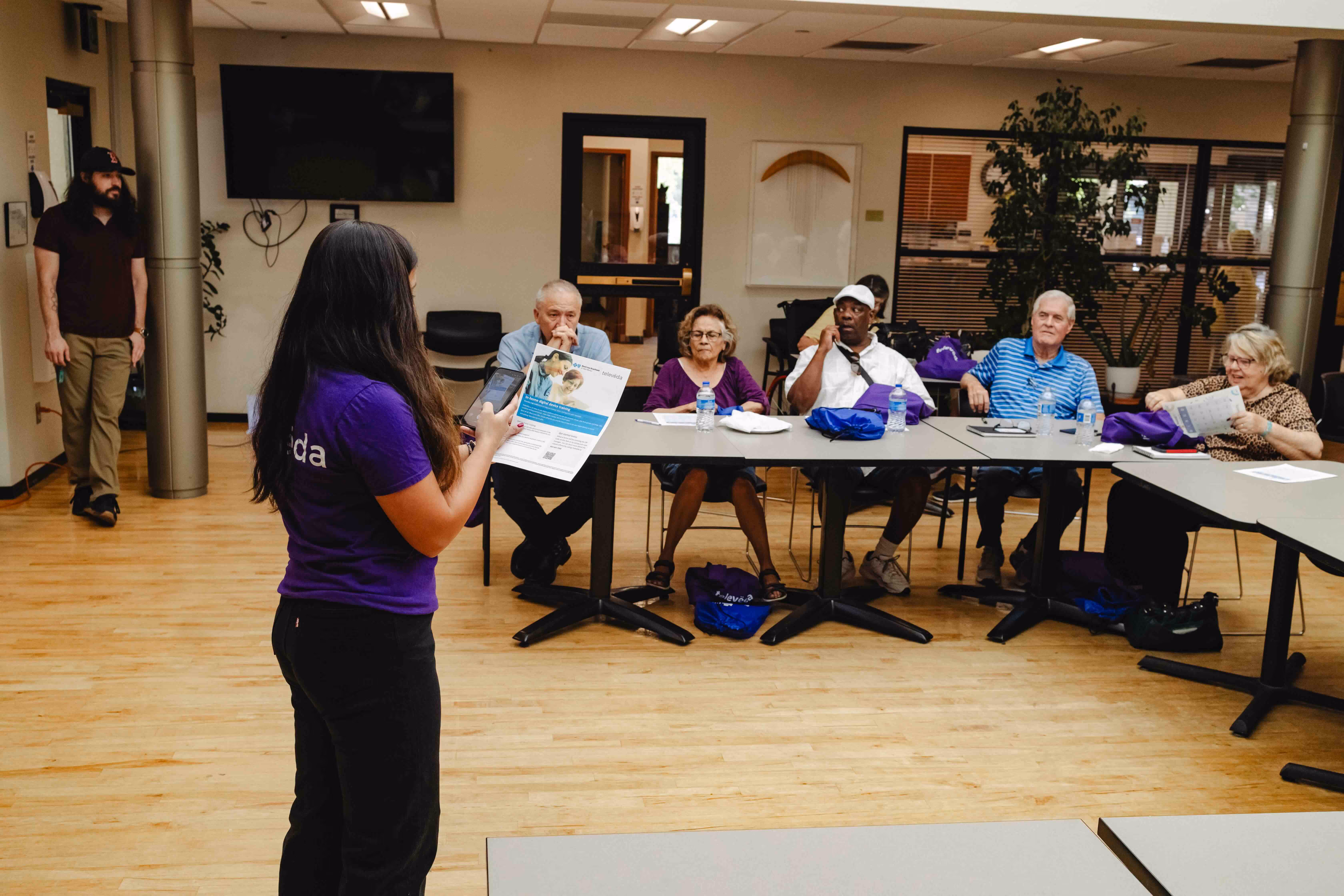A diverse group of adults seated around tables in a classroom setting, engaged in a workshop led by a standing instructor holding a paper.
