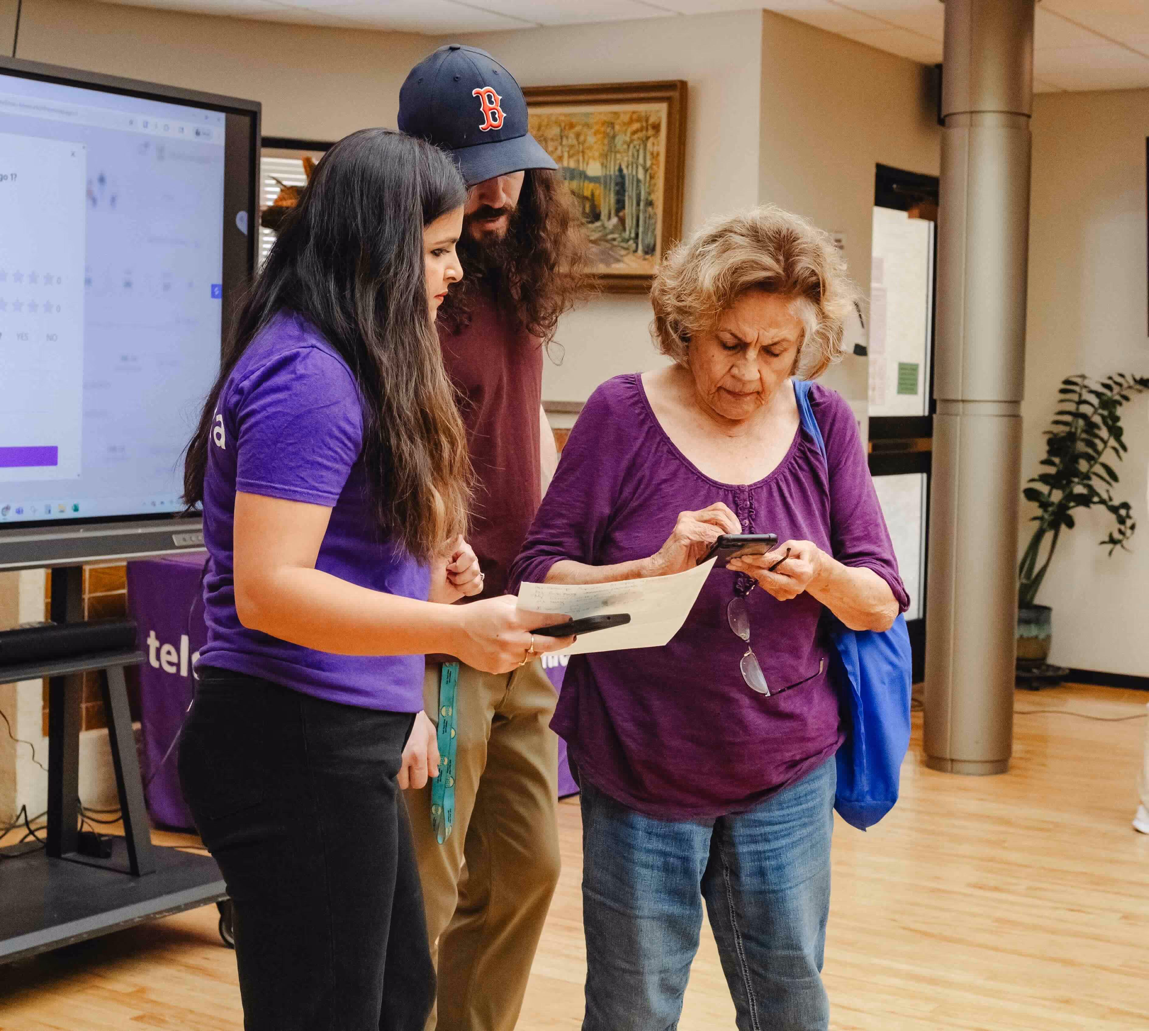 Three people indoors focusing on a smartphone and paper, one elderly woman using the phone while a young woman and man assist her.
