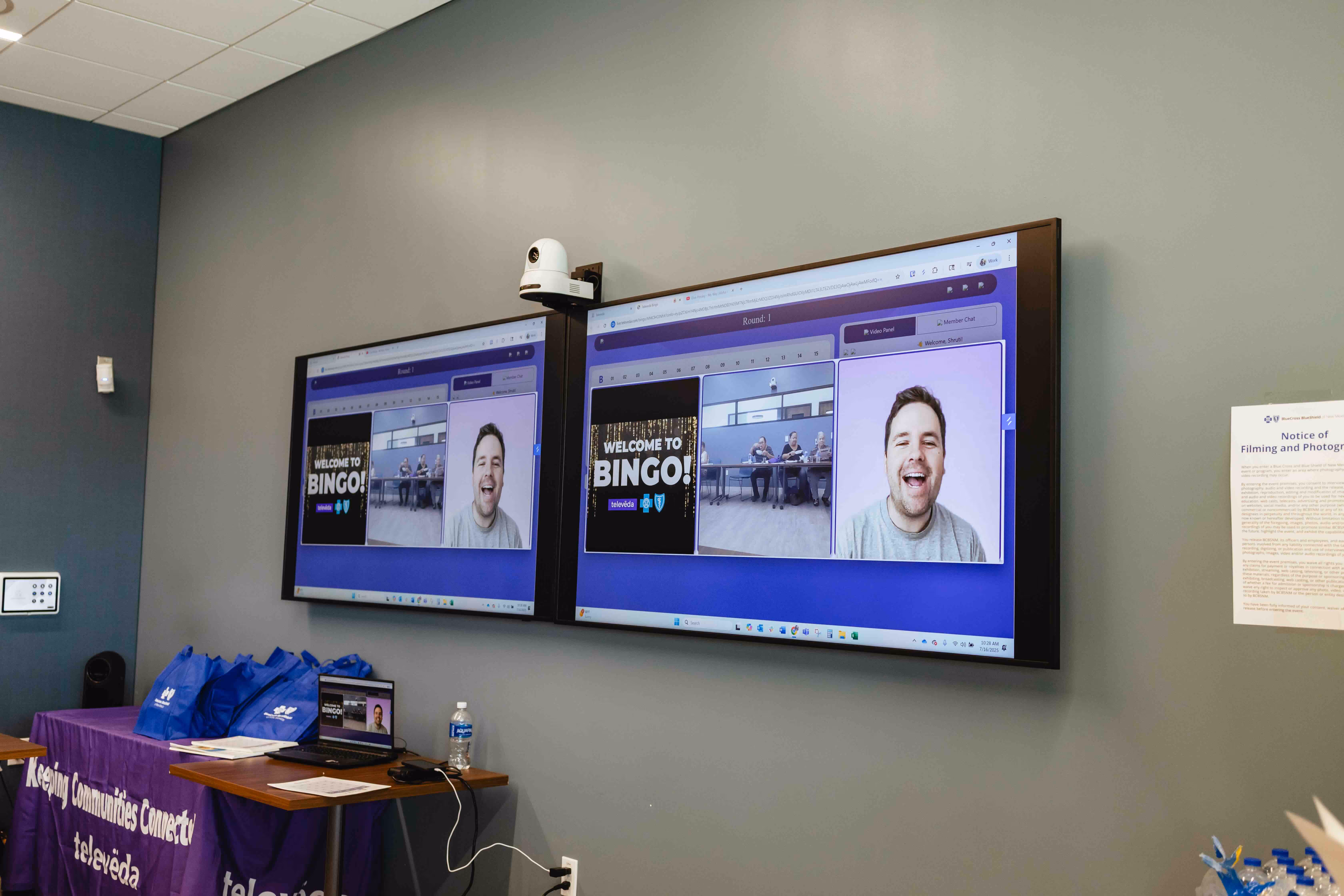 Virtual bingo session displayed on two large monitors mounted on a gray wall, showing a smiling man and people playing bingo remotely.