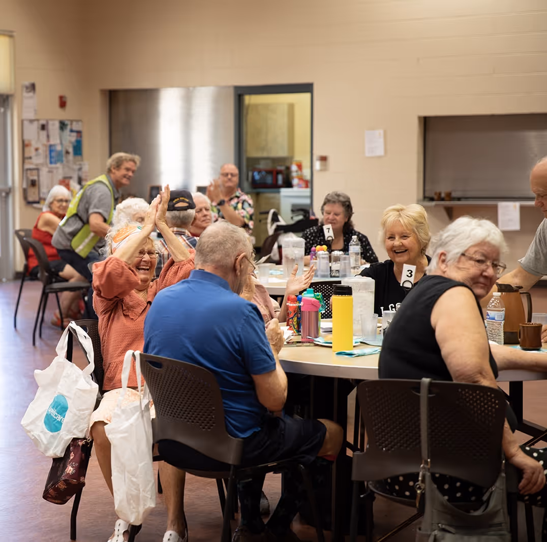 Group of elderly people sitting around tables socializing and smiling in a community center.
