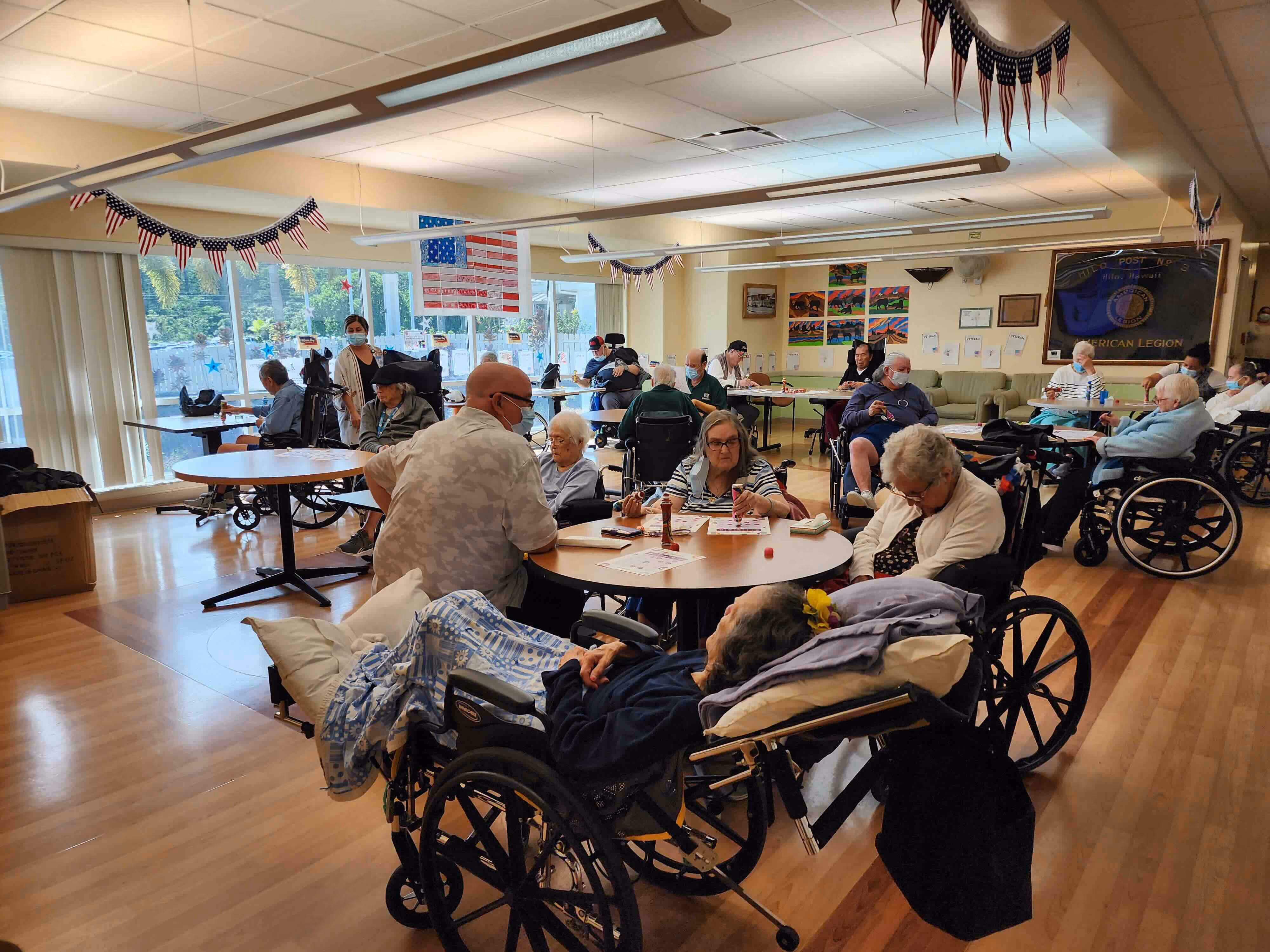Group of elderly people, many in wheelchairs, gathered in a decorated room playing bingo and socializing.