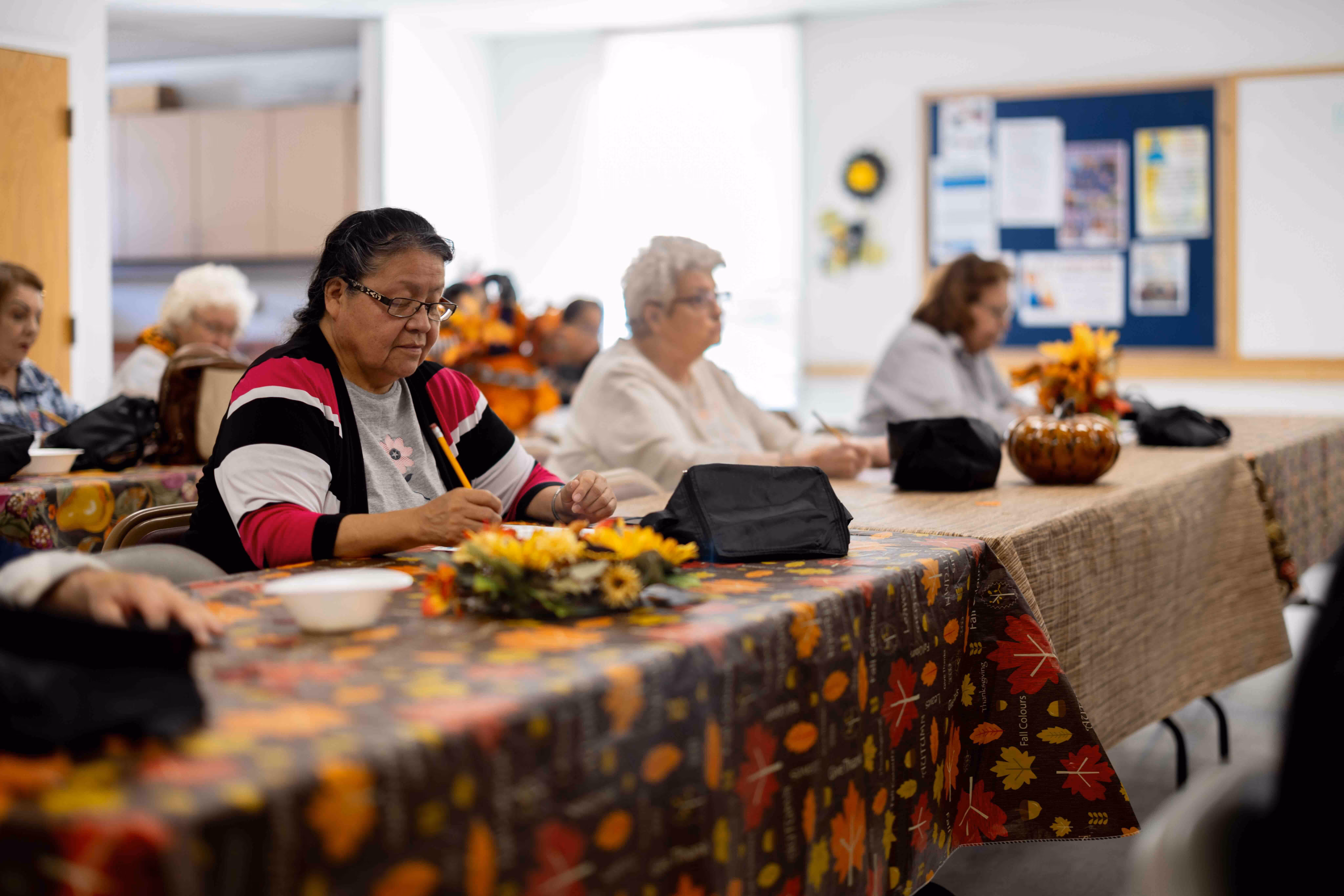 Group of elderly people sitting at decorated tables in a community room, engaging in writing or drawing activities.
