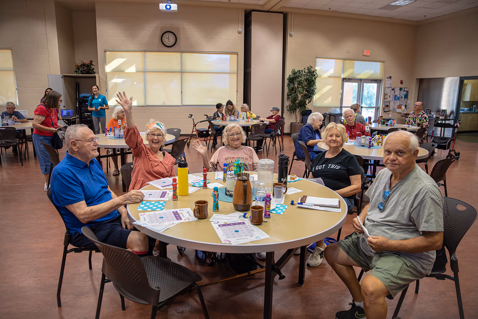 Group of older adults sitting around a table in a community room, playing bingo and smiling, with some waving at the camera.