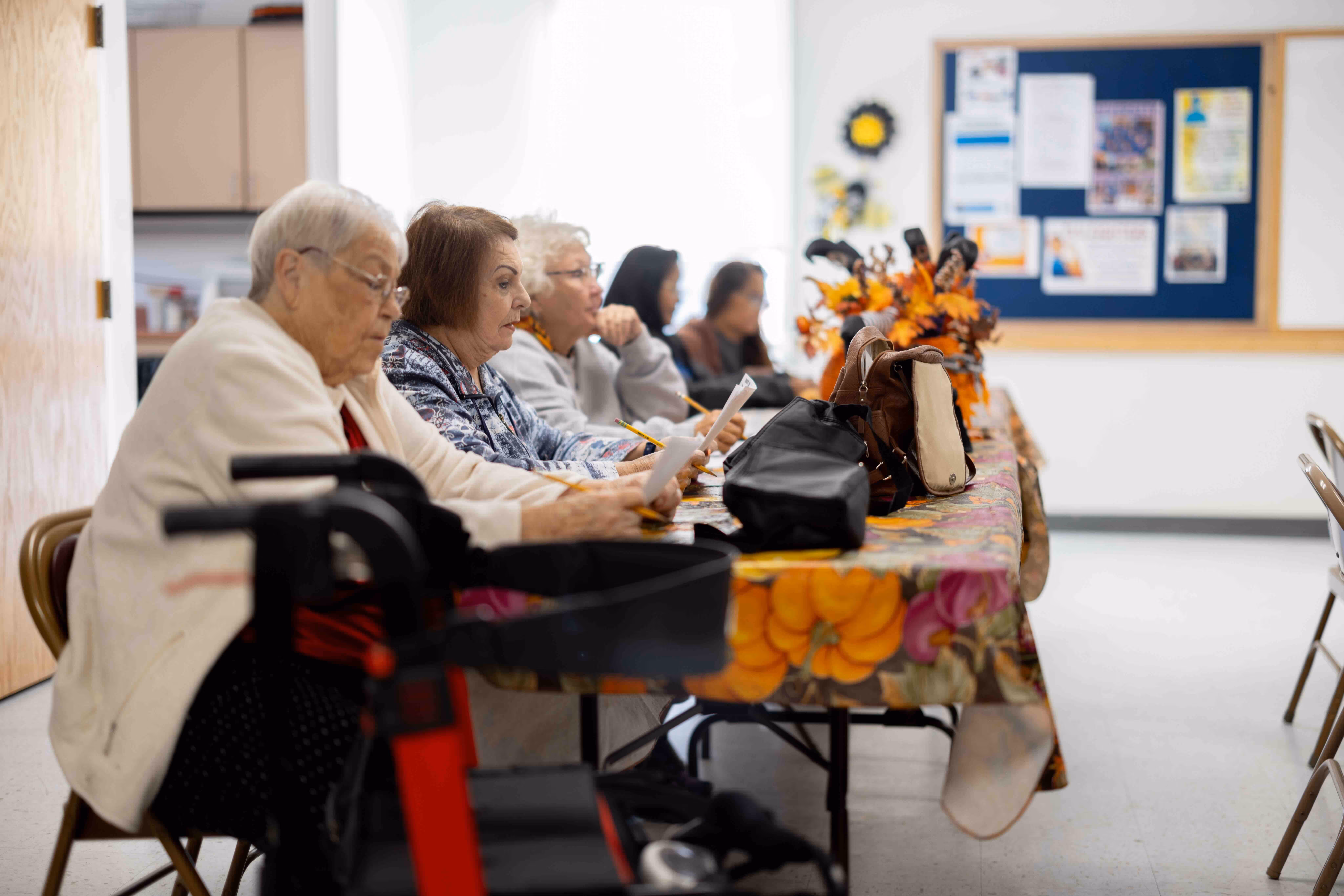 A group of elderly individuals sitting at a long table with a floral tablecloth, engaged in writing or reading papers.