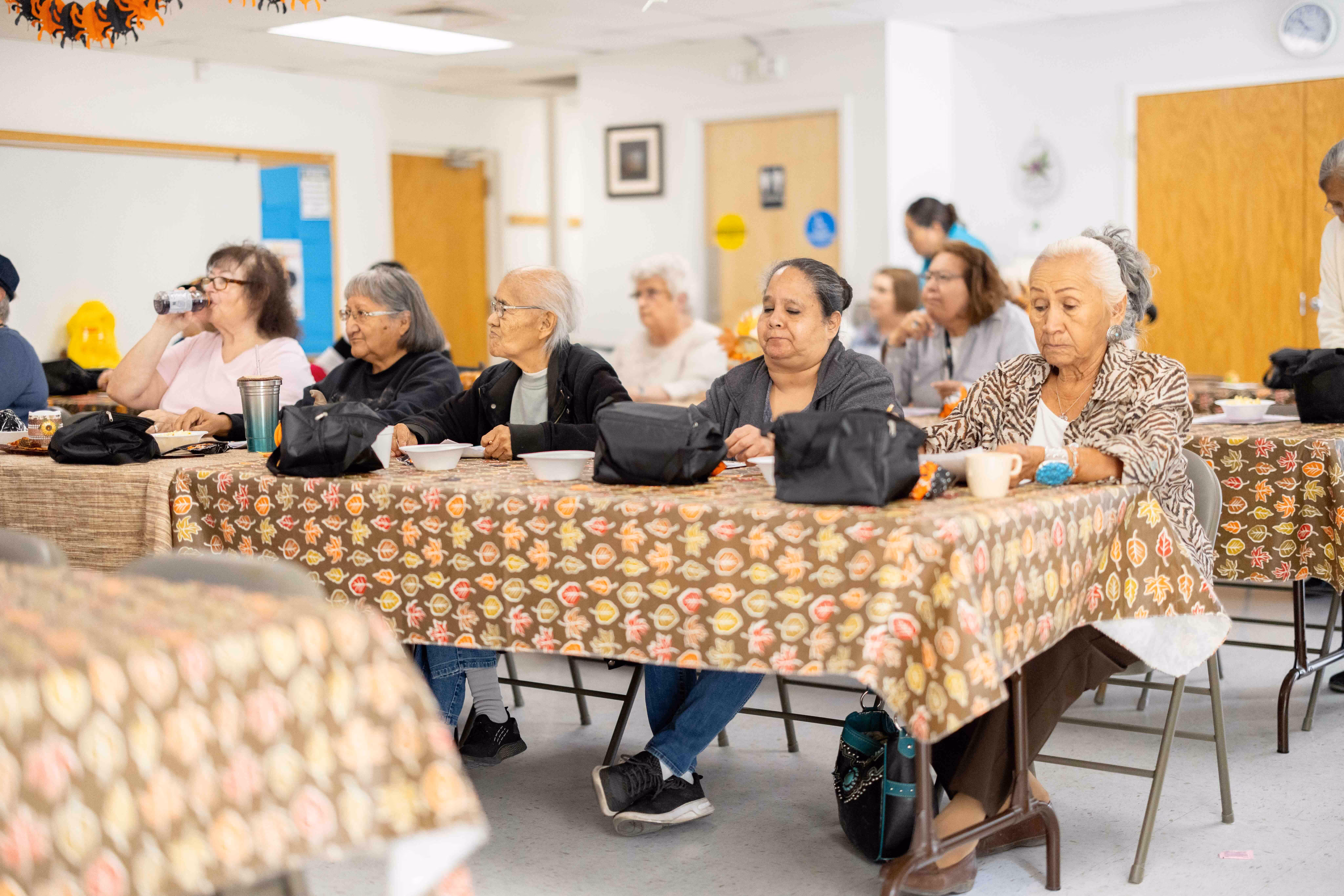 Group of elderly people sitting at tables with autumn leaf patterned tablecloths, eating and drinking in a community center.