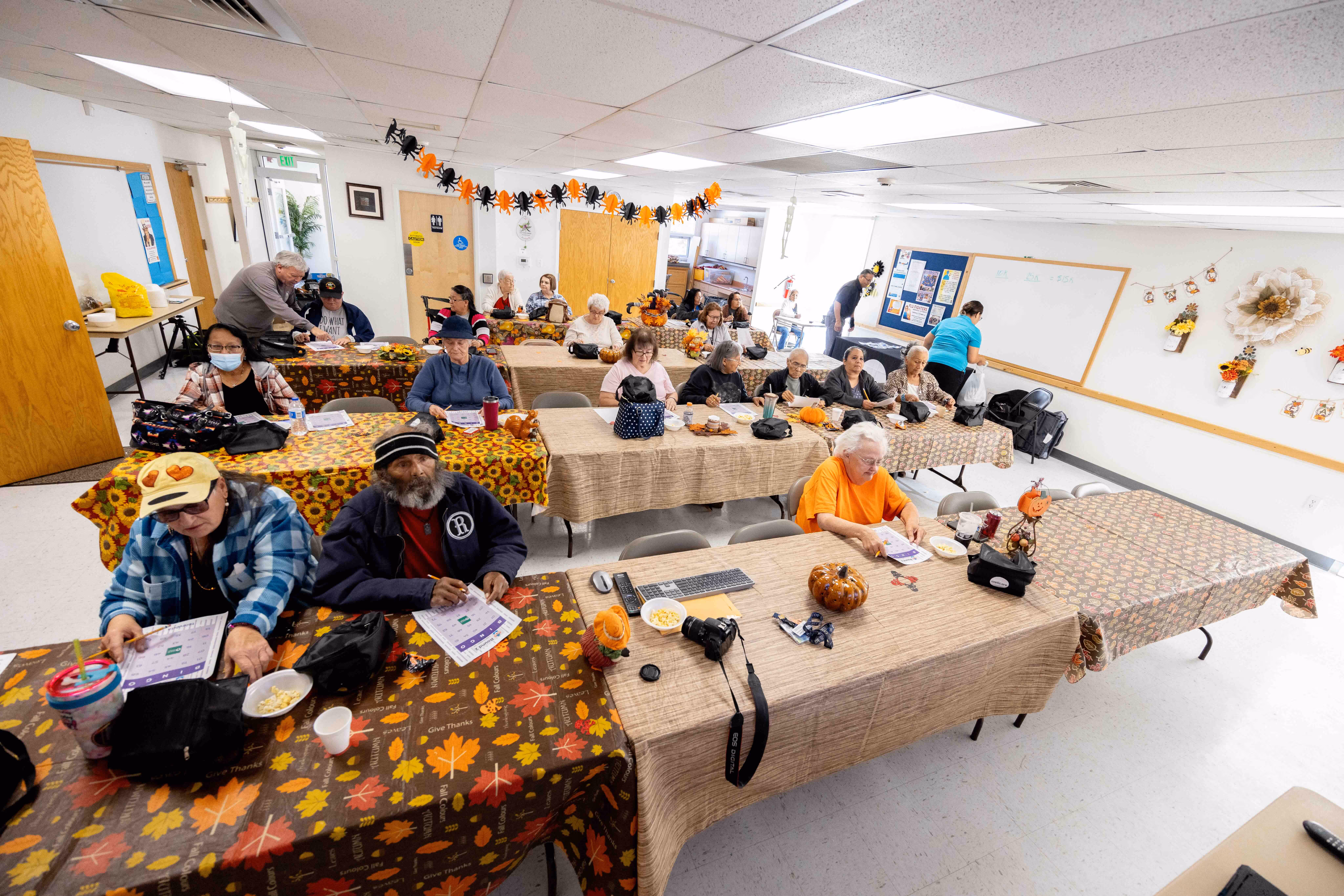 Group of elderly people sitting at decorated tables in a community room, engaging in an activity with papers and snacks.