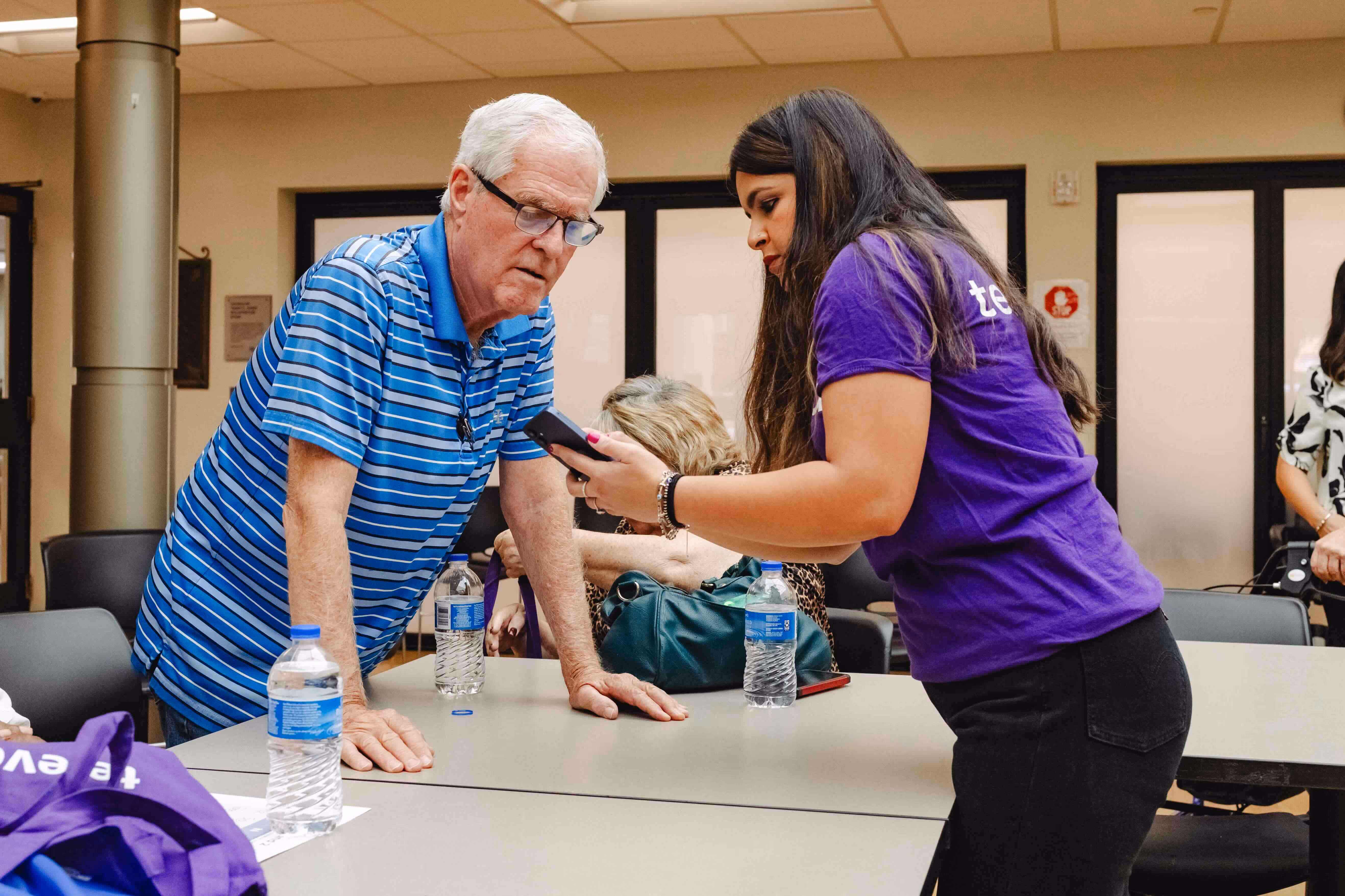 A woman in a purple shirt shows a smartphone screen to an elderly man in a blue striped shirt at a table with water bottles.
