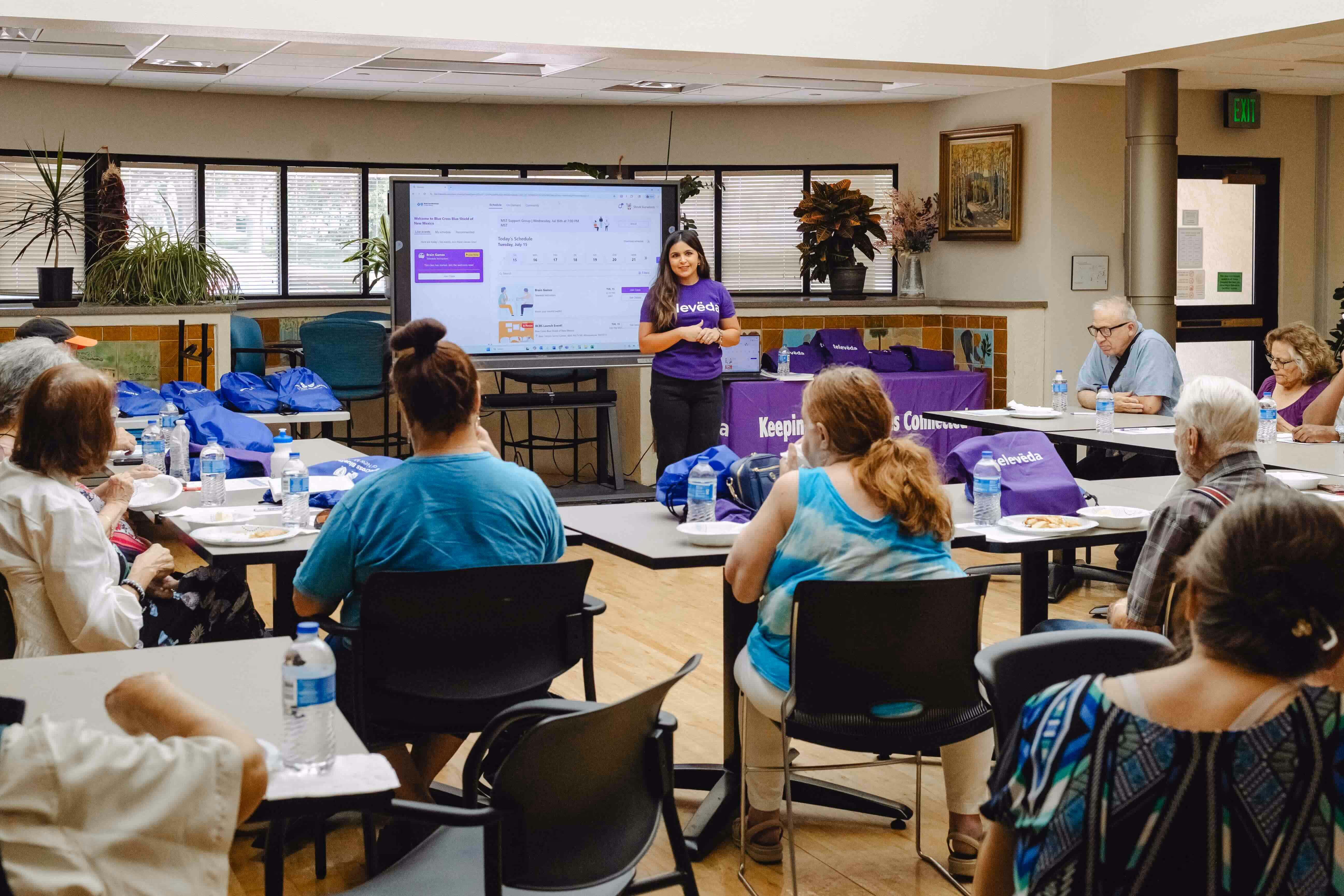 Woman in a purple shirt giving a presentation to a seated group of people in a community room.