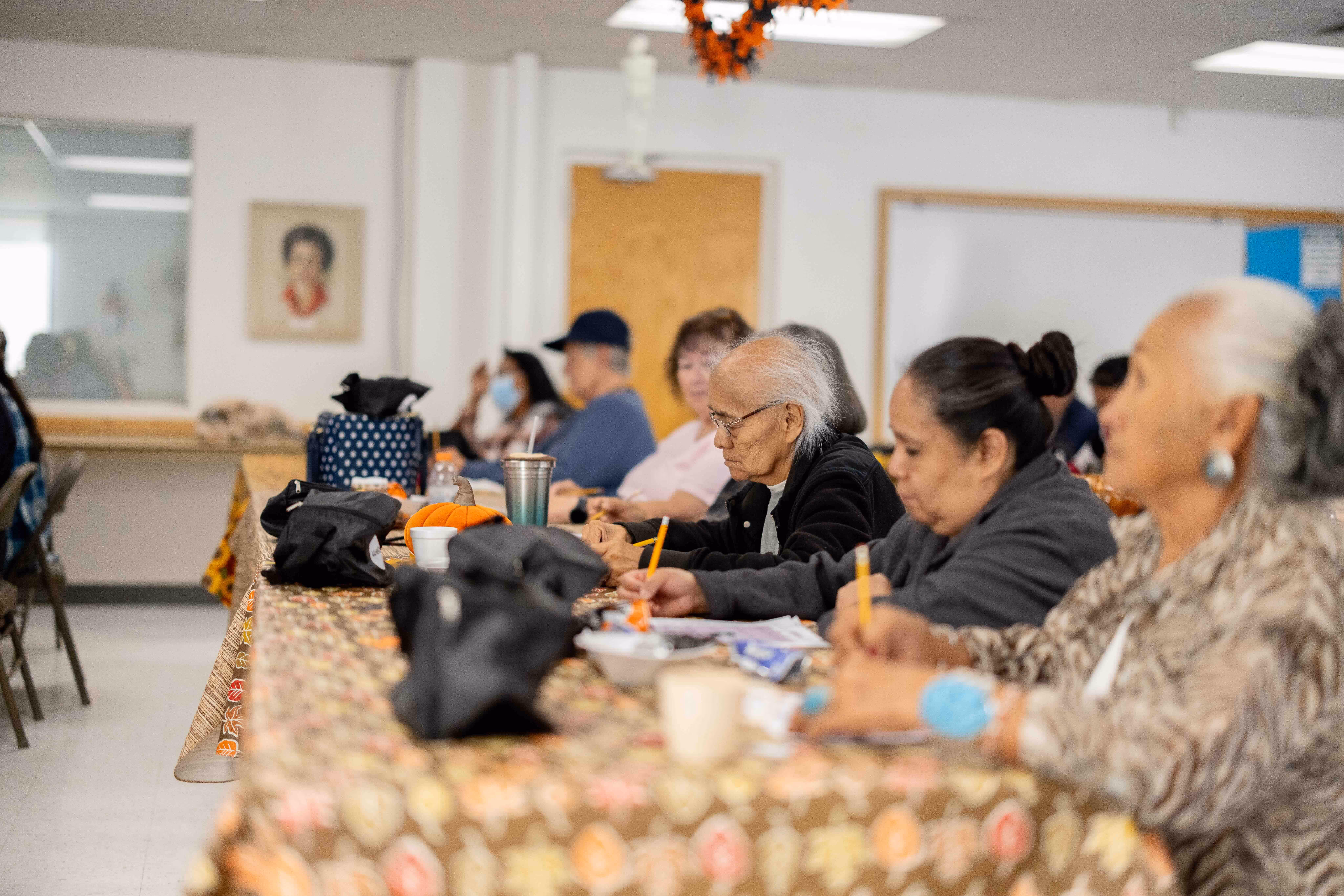 Elderly people sitting at a long table with floral tablecloth, engaged in writing or drawing activities.