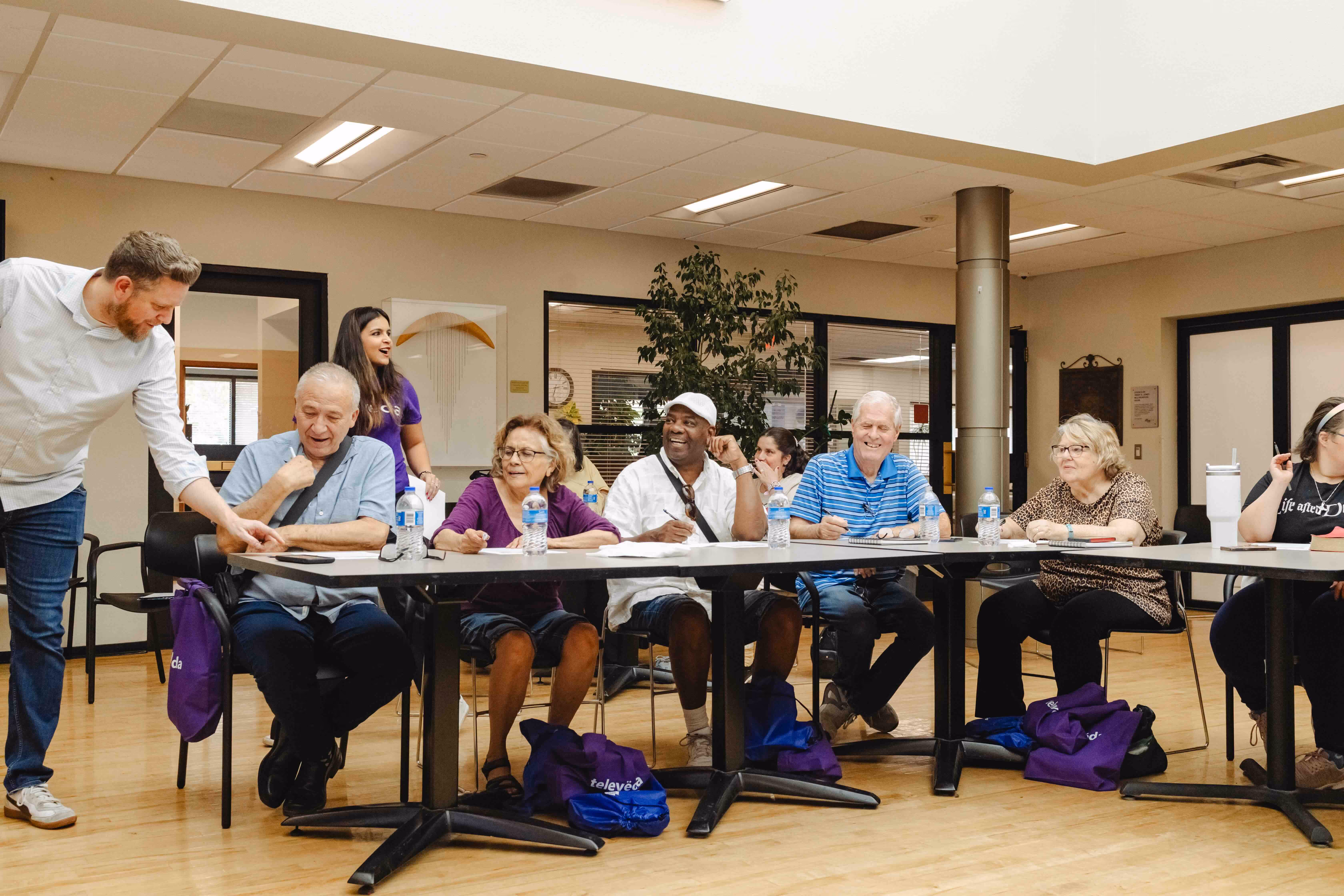 A diverse group of adults sitting around tables in a classroom setting, engaging with a standing instructor.