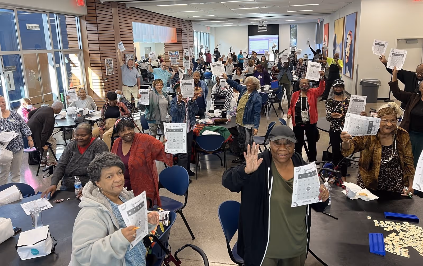 A large group of diverse older adults in a community room holding up bingo cards, smiling and celebrating.