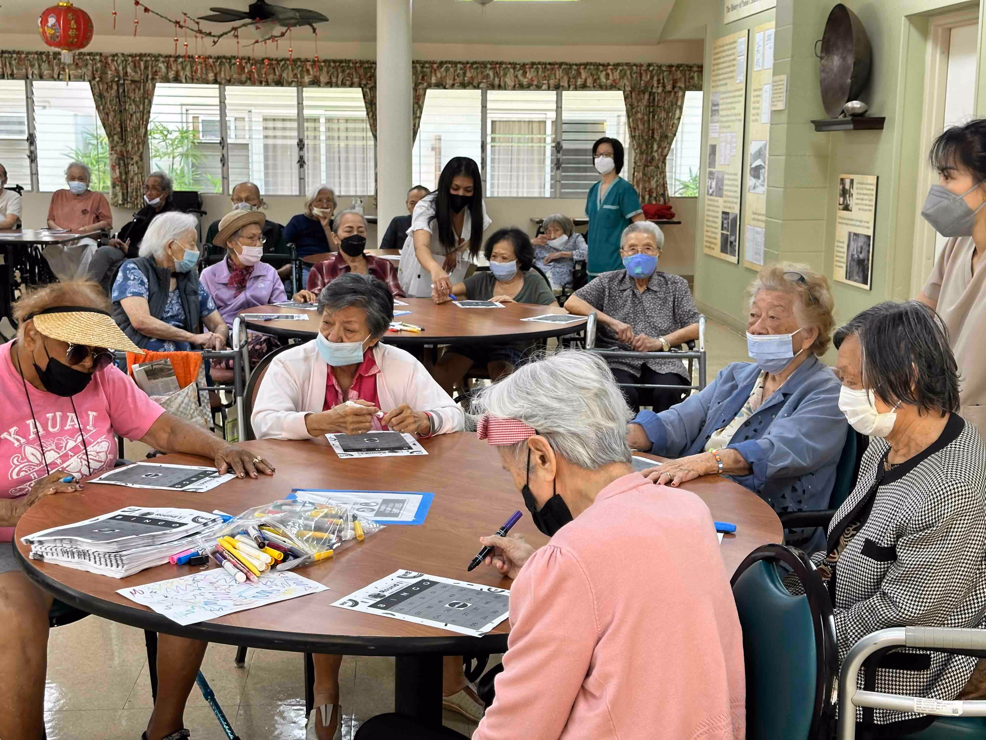 Group of elderly people wearing masks sitting around tables doing activities with pens and papers in a communal room with caregivers assisting.