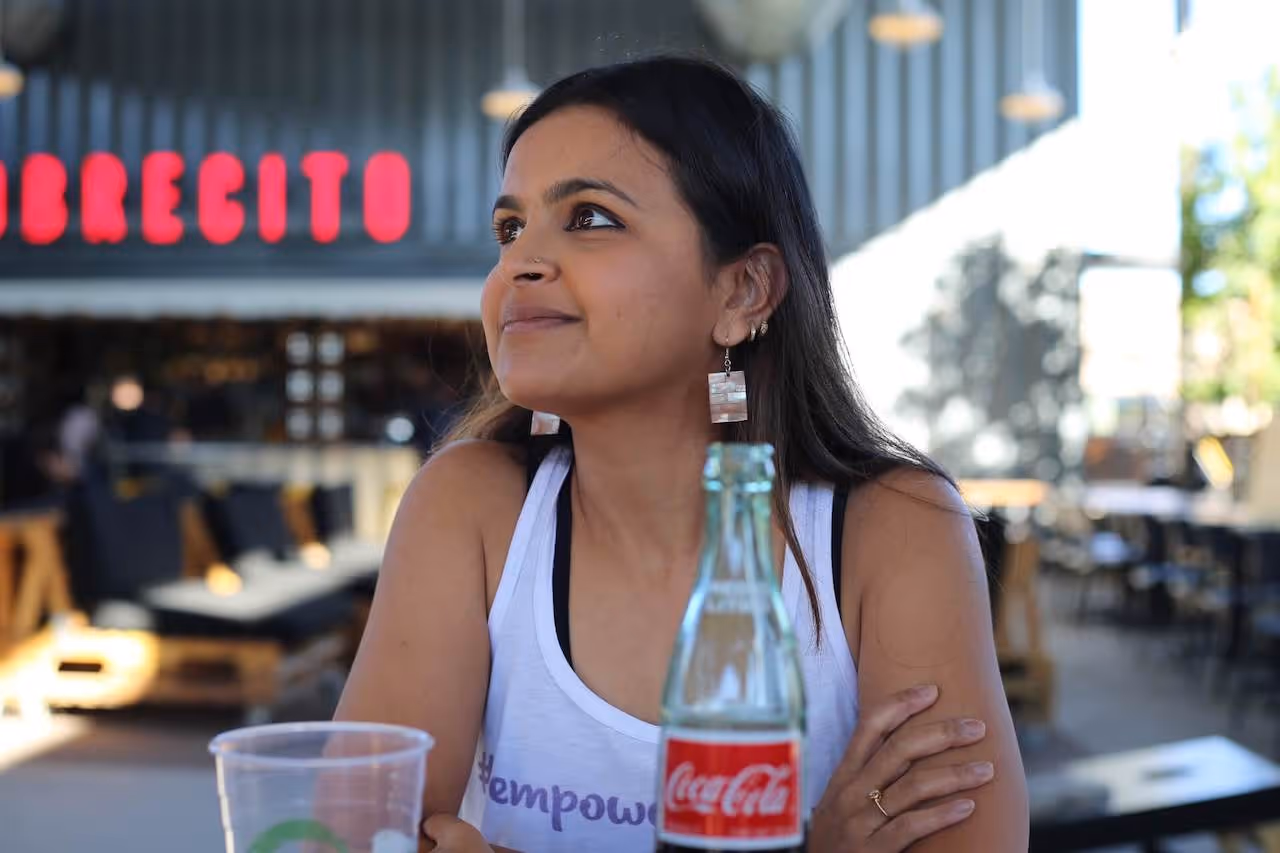 Woman with dark hair wearing a white tank top and rectangular earrings sitting at an outdoor table with a Coca-Cola bottle and plastic cup in front of her.