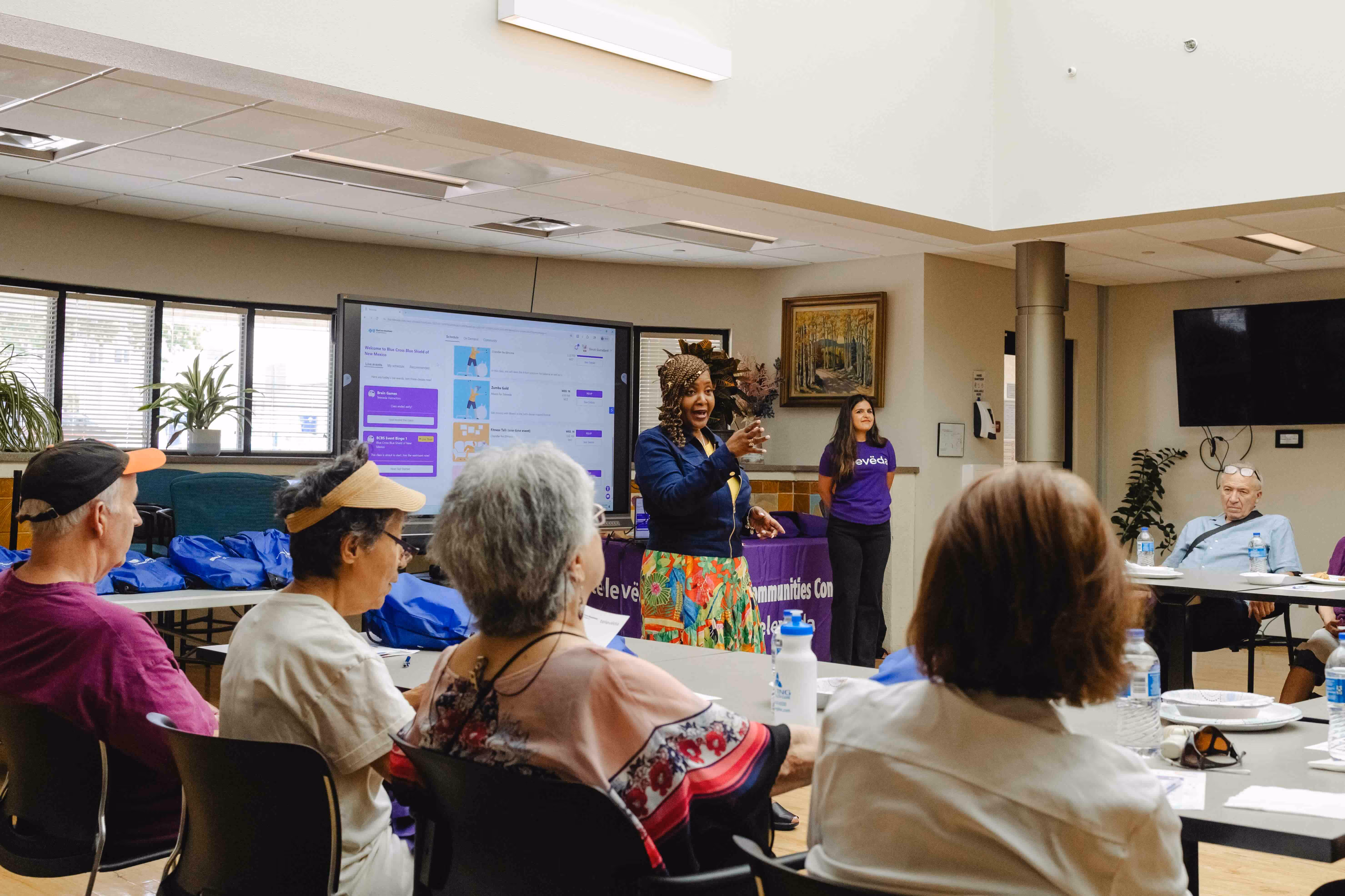 A woman speaks to a seated group of older adults in a community room with a large screen displaying a website behind her.