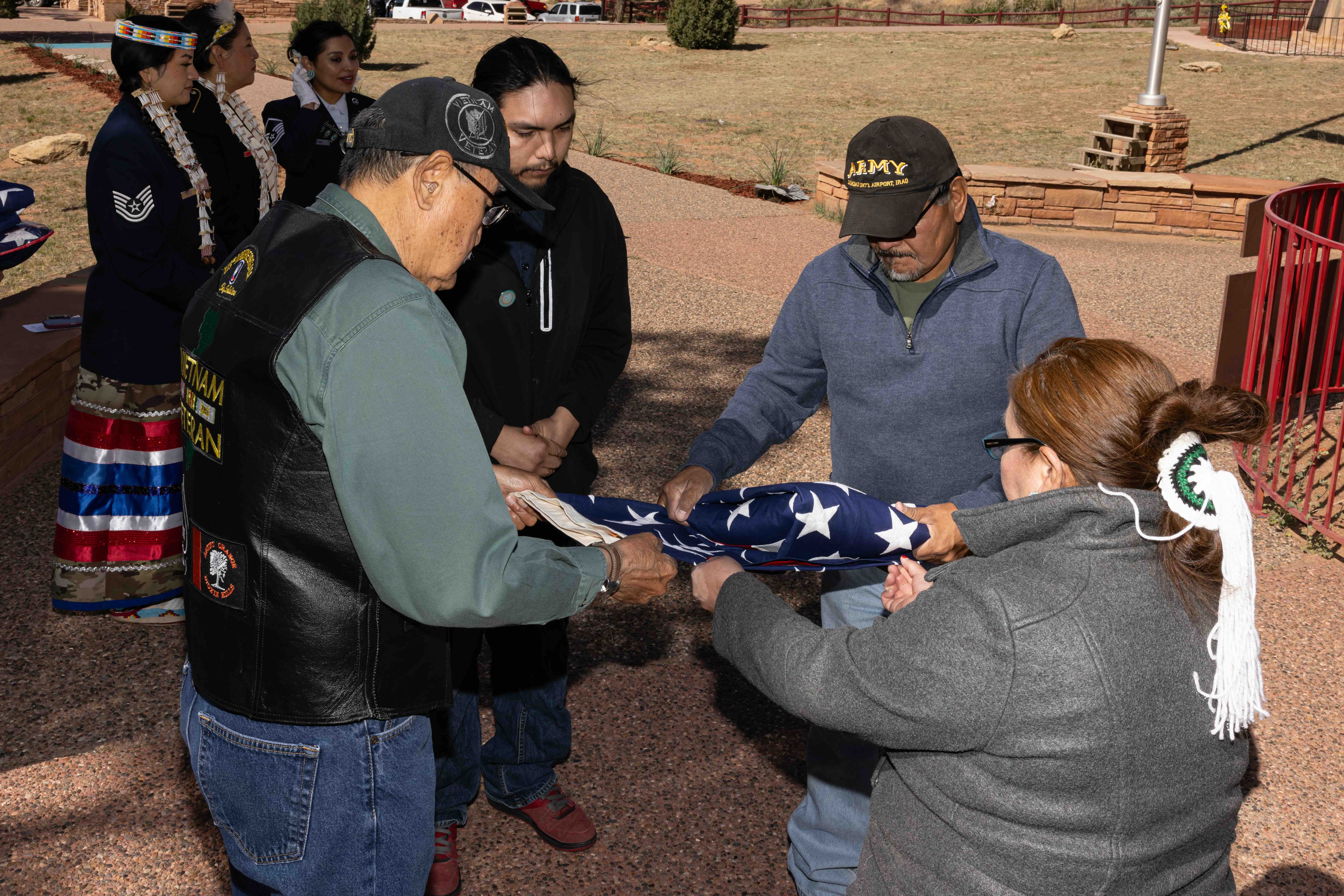 Four people carefully folding an American flag outdoors, with three women dressed in traditional Native American attire standing in the background.