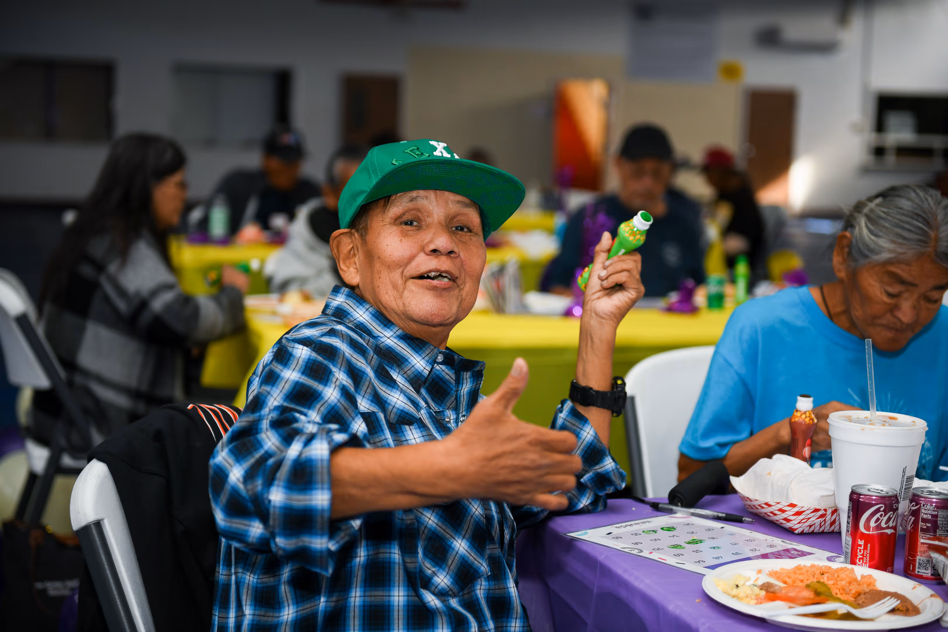 Elderly person in a green cap and blue plaid shirt giving a thumbs-up while holding a green bingo dauber, seated at a table with food and drinks during a group event.