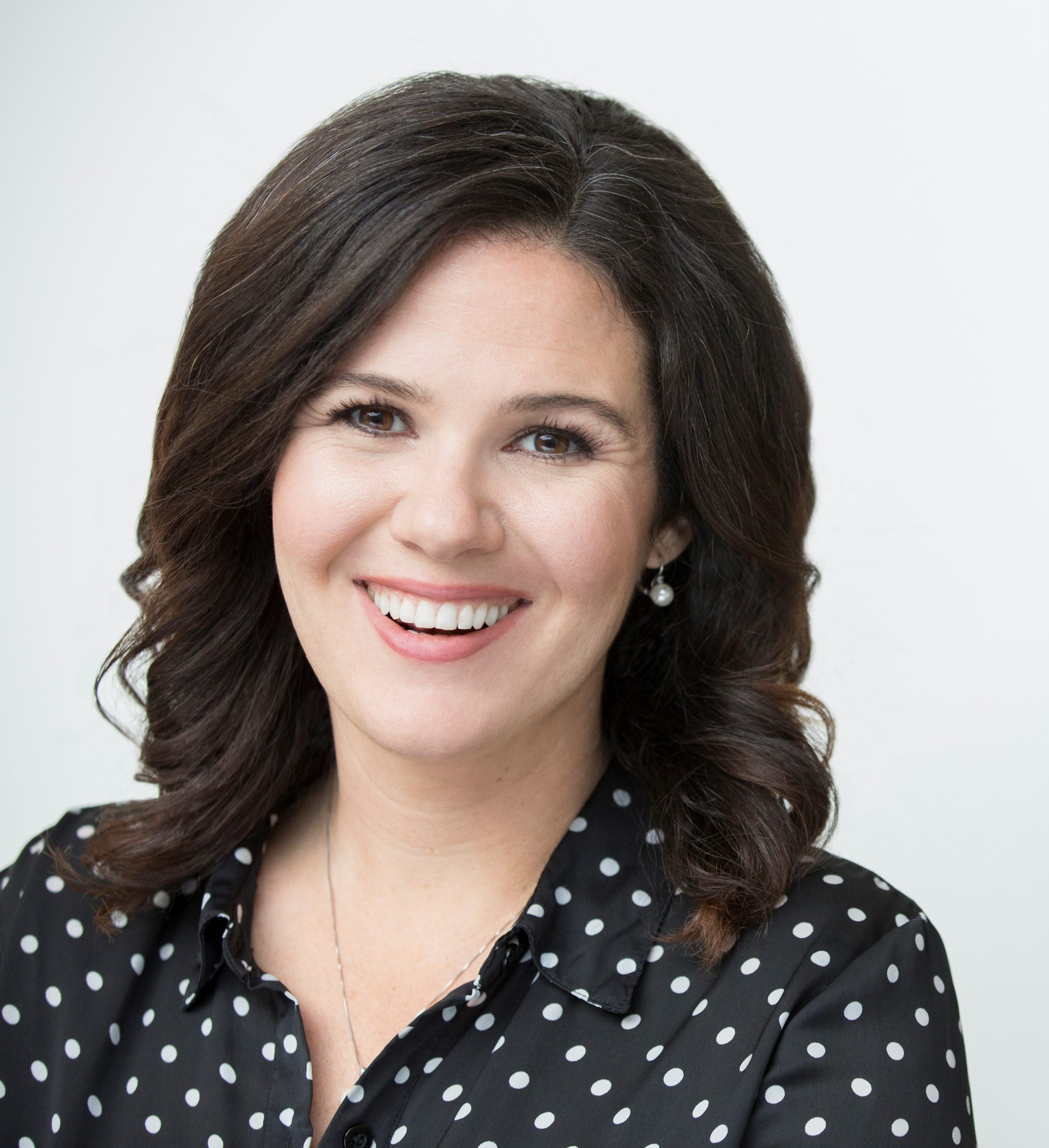 Smiling woman with shoulder-length dark hair wearing a black polka dot blouse and pearl earrings in front of a plain white background.