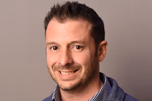 Smiling man with short dark hair wearing a blue jacket and checkered shirt, against a plain background.