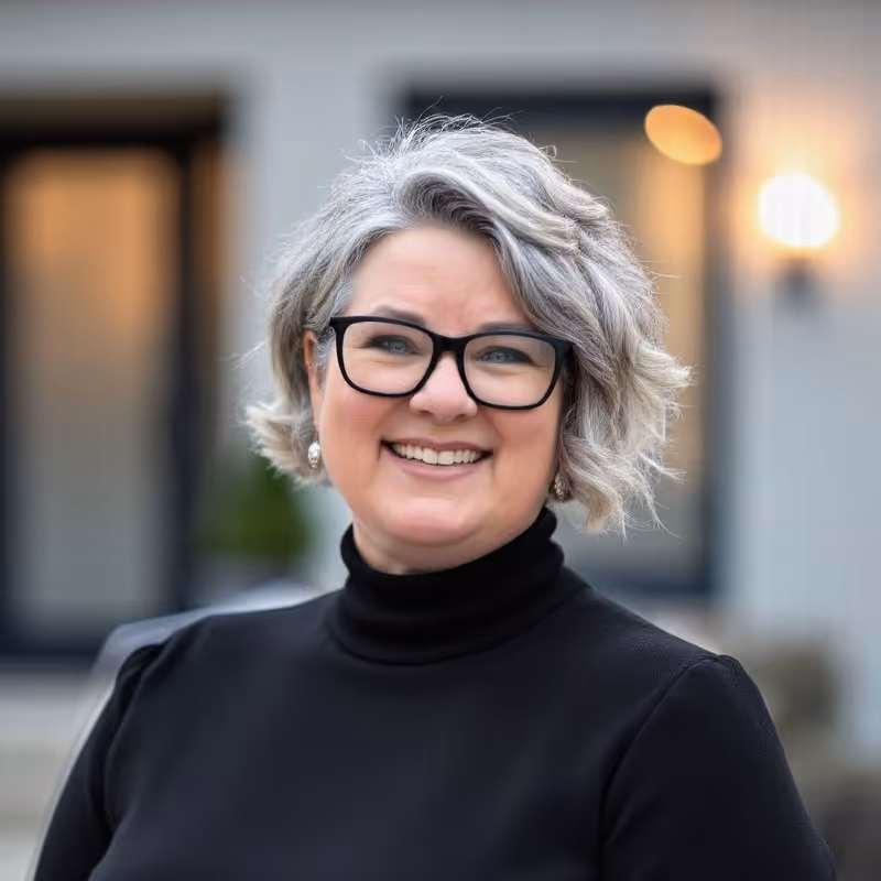 Smiling middle-aged woman with short gray hair, black glasses, and a black turtleneck sweater, standing outdoors.