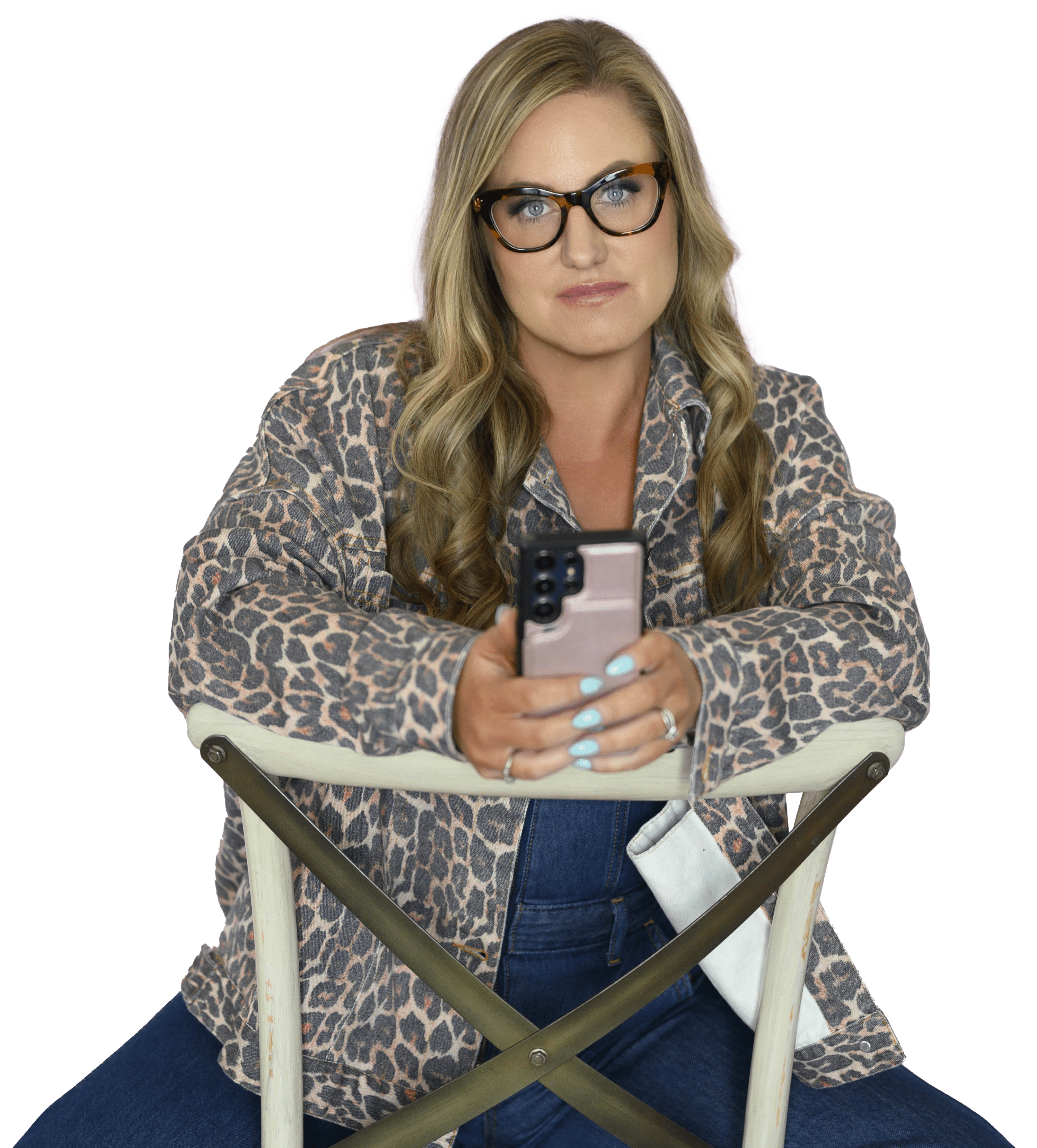 Woman with long blonde hair and glasses sitting backwards on a chair, holding a smartphone and wearing a leopard print jacket.