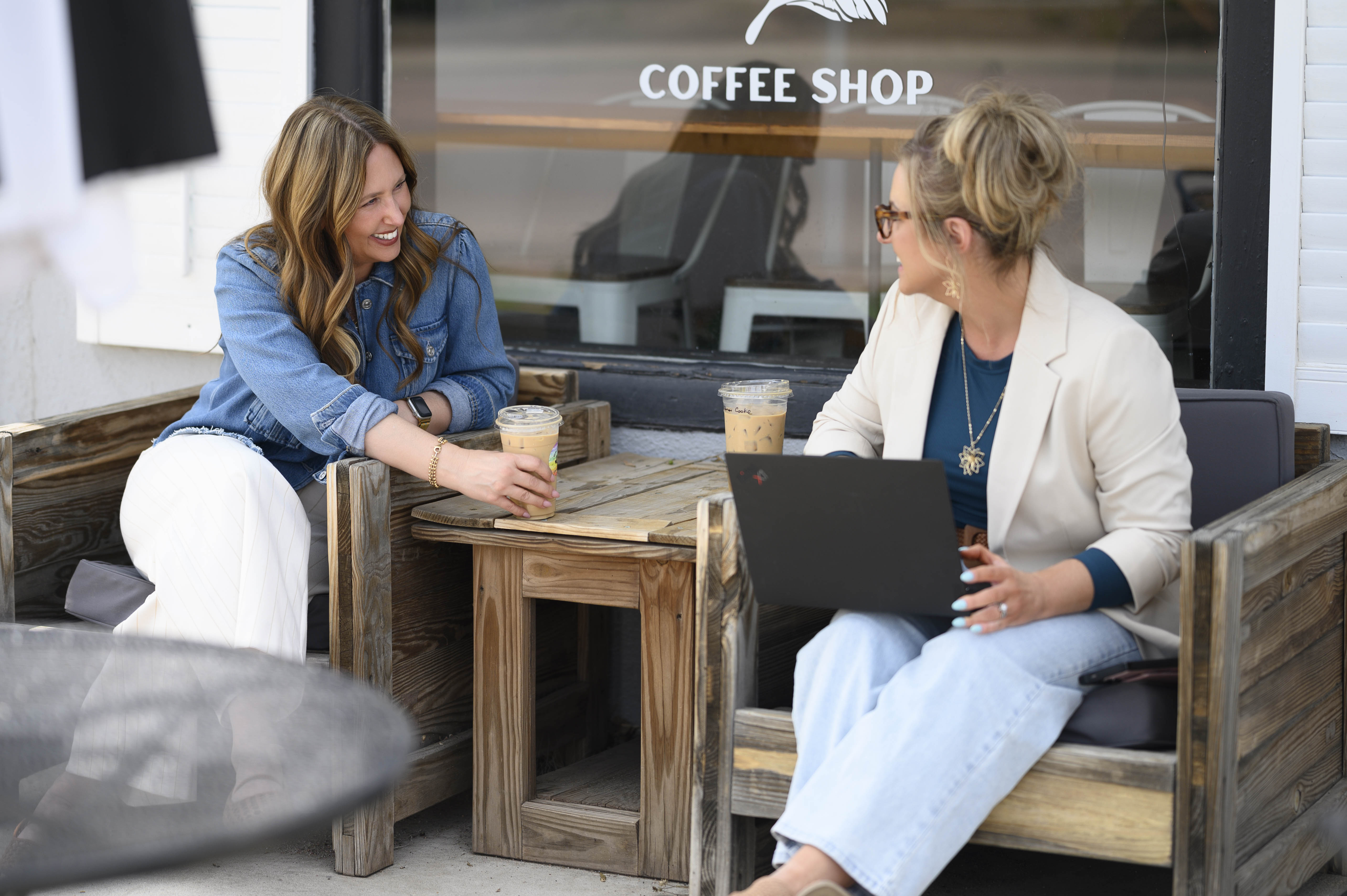 Two women sitting on wooden chairs outside a coffee shop, one holding iced coffee and the other using a laptop.