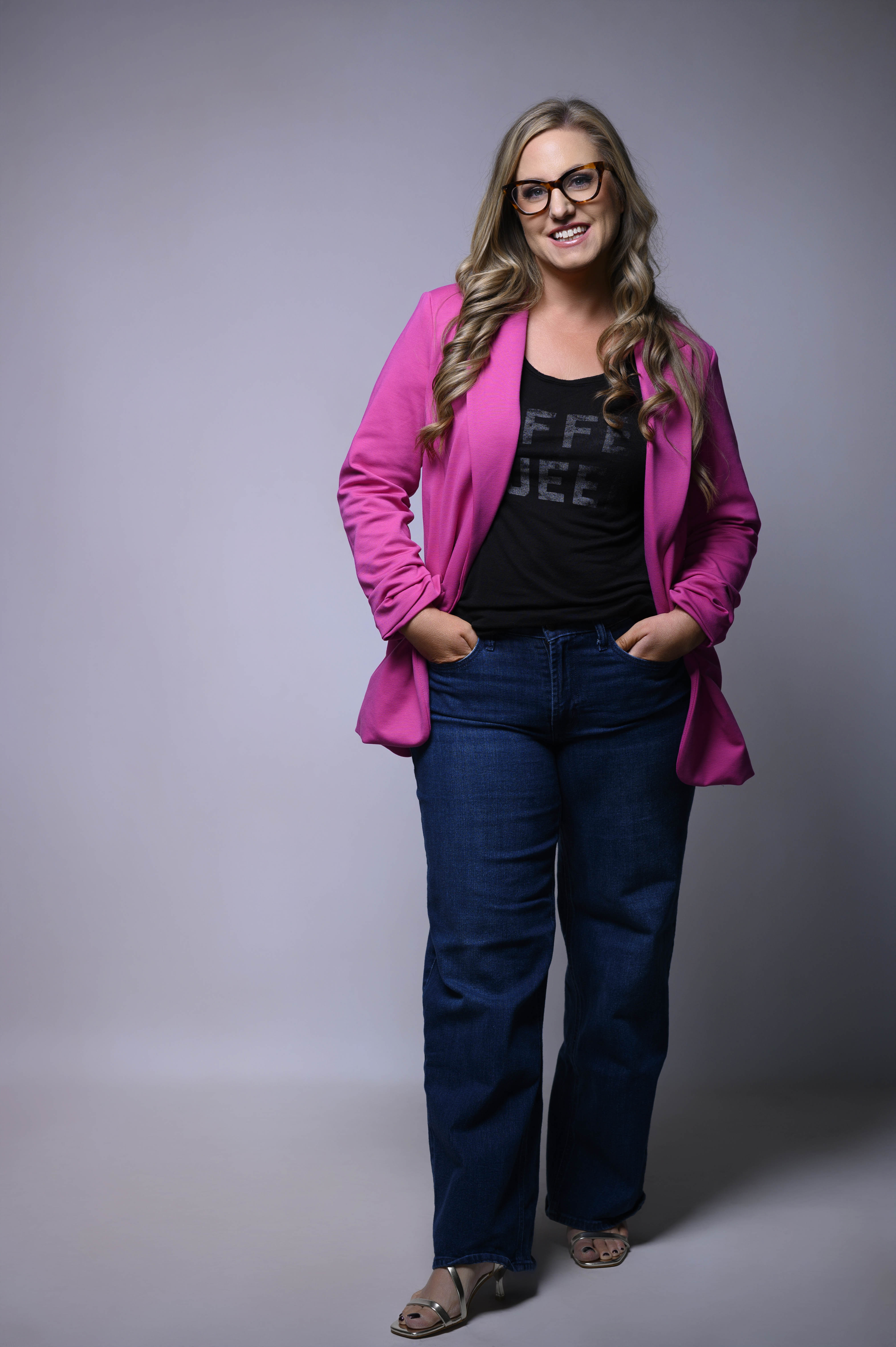 Smiling woman with long wavy blonde hair wearing glasses, a bright pink blazer, black top, and blue jeans, standing with hands in pockets against a plain gray background.