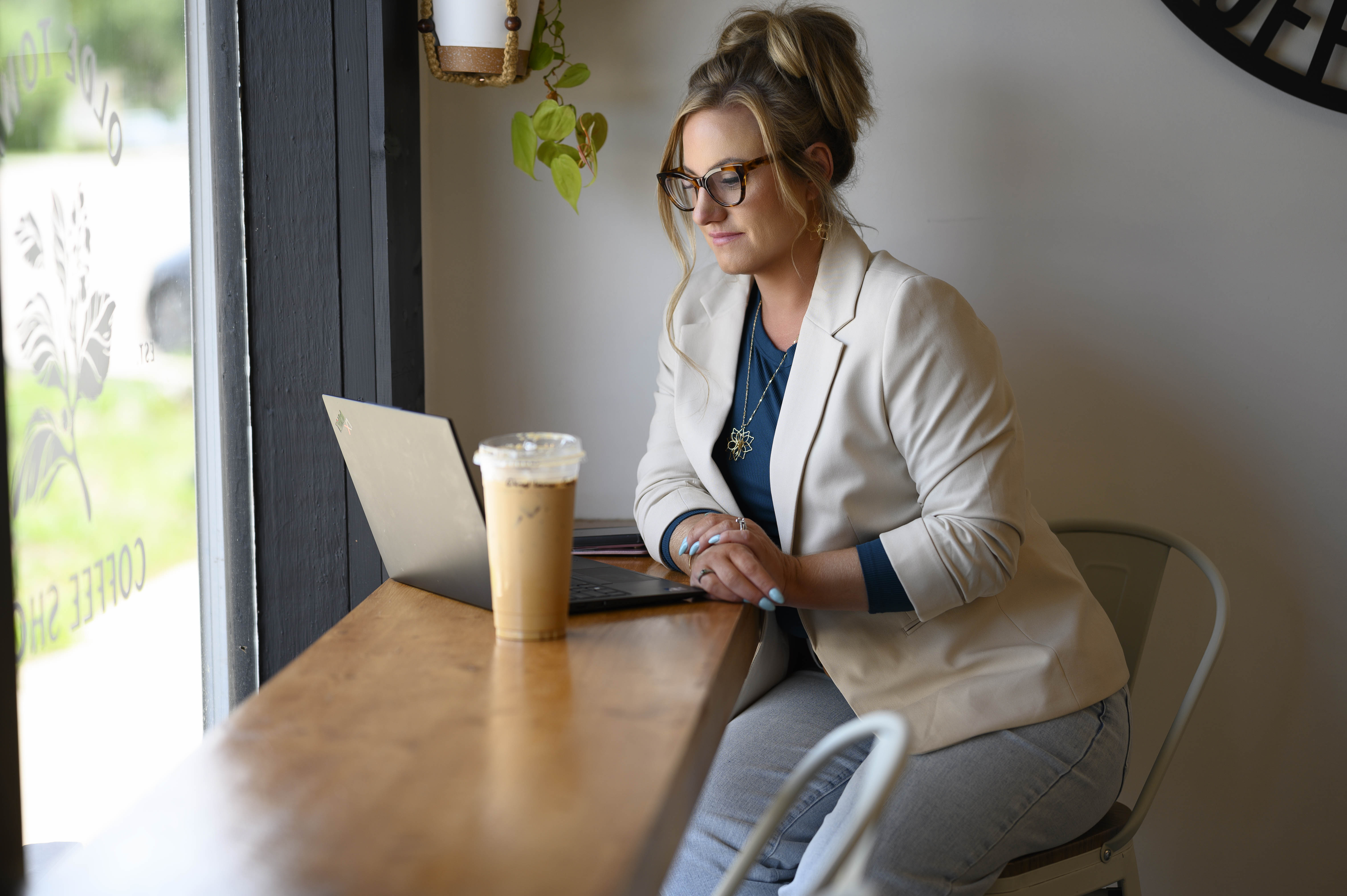 Woman wearing glasses and a beige blazer sitting at a coffee shop counter, working on a laptop with an iced coffee nearby.