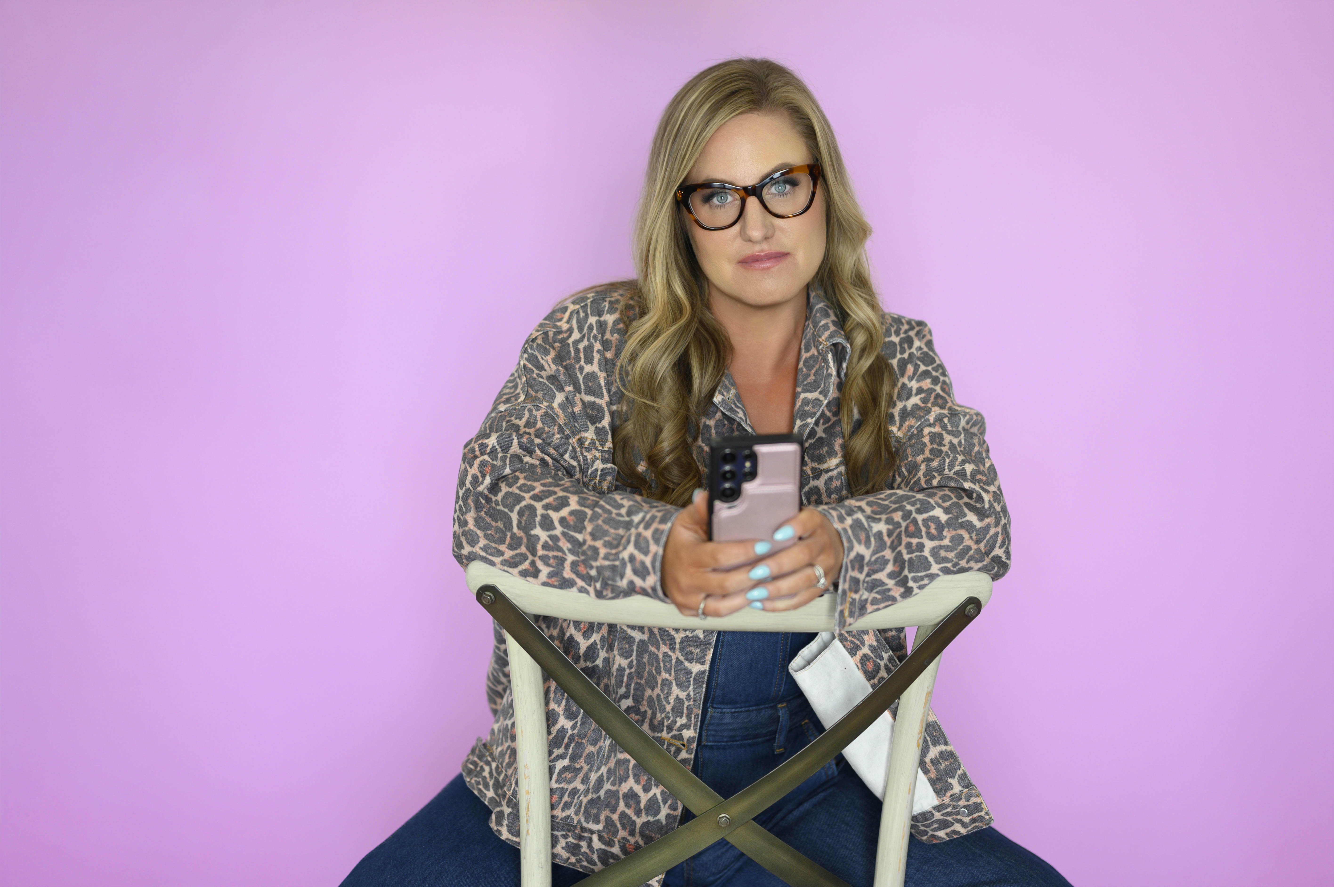 Woman with long blonde hair and glasses sitting backwards on a chair, holding a smartphone against a pink background.