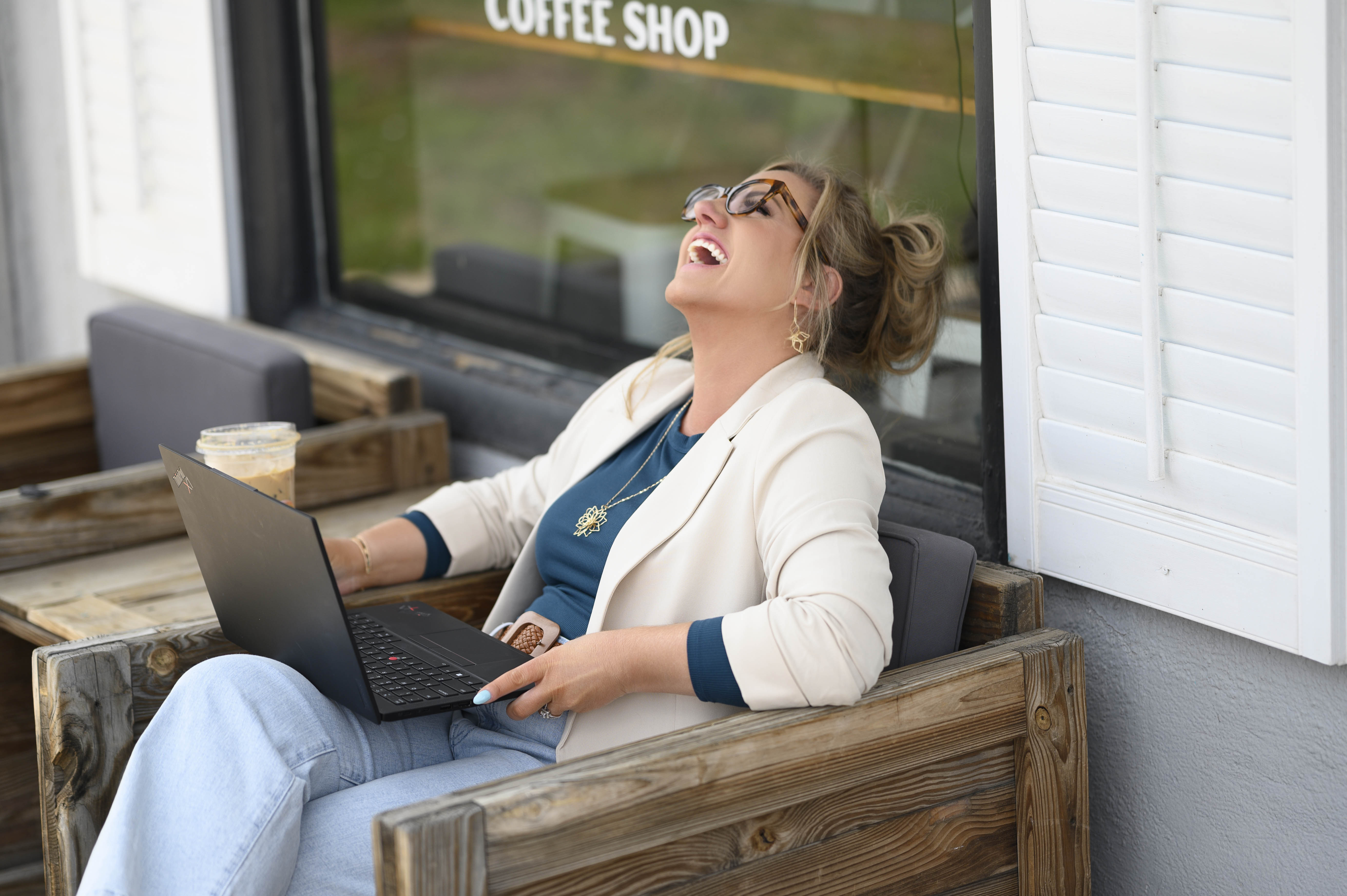 Woman wearing glasses and a white blazer laughing while sitting on a wooden outdoor chair with a laptop and iced coffee at a coffee shop.