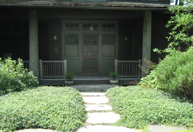 Front view of a dark shingled Craftsman-style home porch. Square stepping stones are set in groundcover leading to the entrance.