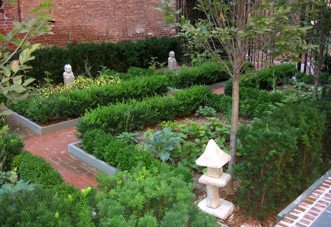 Formal urban garden beds bordered by brick paths, featuring boxwood hedges, a stone lantern, and small statues.