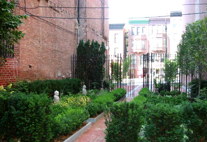 Brick pathway leading through formal hedged garden beds and iron gate to a row of brownstone buildings across the street.