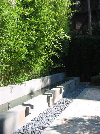 Modern urban water feature with bamboo screen, smooth concrete troughs, and a channel filled with dark river stone.
