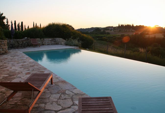 Infinity edge swimming pool with natural stone patio and cypress trees, overlooking the Tuscan hills at sunset.