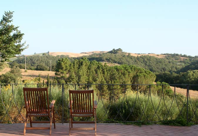 Two wooden chairs on a brick patio, overlooking a vast, lush valley of rolling Tuscan farmland and trees.