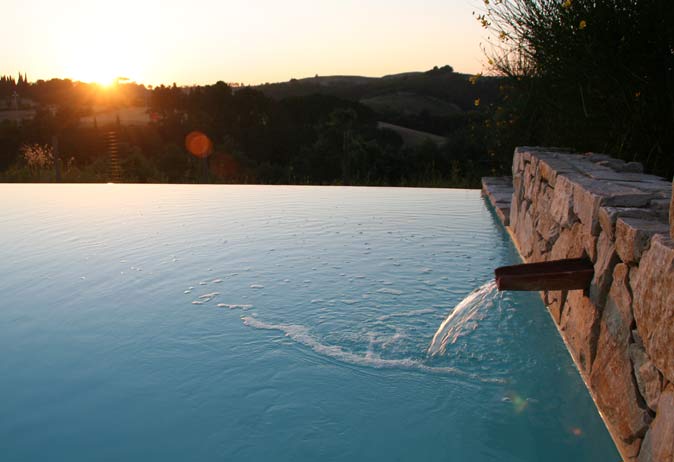 Close-up of the infinity edge pool with a rustic stone wall, copper spout, and sunset over the rolling Tuscan landscape.