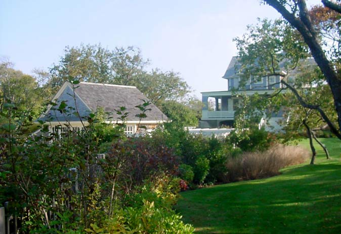 View across a tiered garden bed toward a shingled house and a smaller outbuilding, separated by a white fence.
