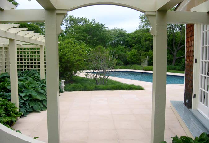View through a white pergola of a pool patio with pale paving stones, lush grass beds, and surrounding trees.