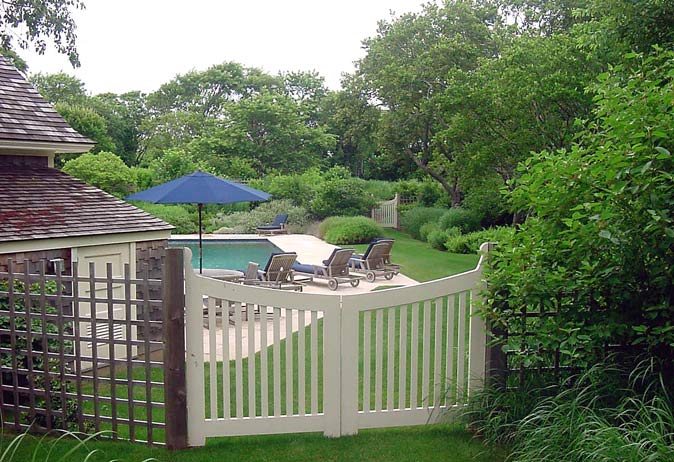 White wooden double gate with decorative tops opening onto a pool deck with lounge chairs and pool house.