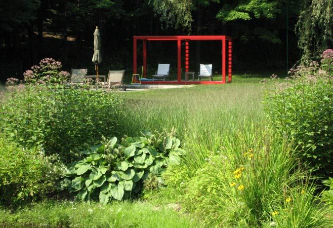 Pond-side view of a red modern structure and lounge chairs set in a manicured lawn, framed by tall perennial plantings.