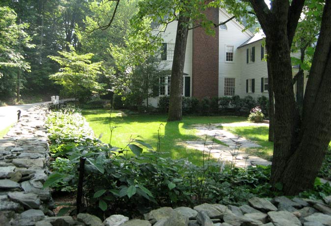 Traditional white house with stone walls and mature shade trees, bordering a stone path and lush, sunlit lawn.
