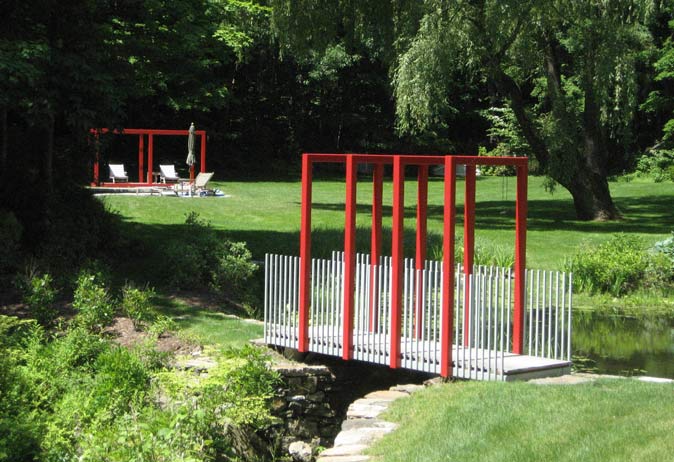 Modern red and white bridge over a stream, leading to a red open-air pavilion and lounge area under a large willow tree.